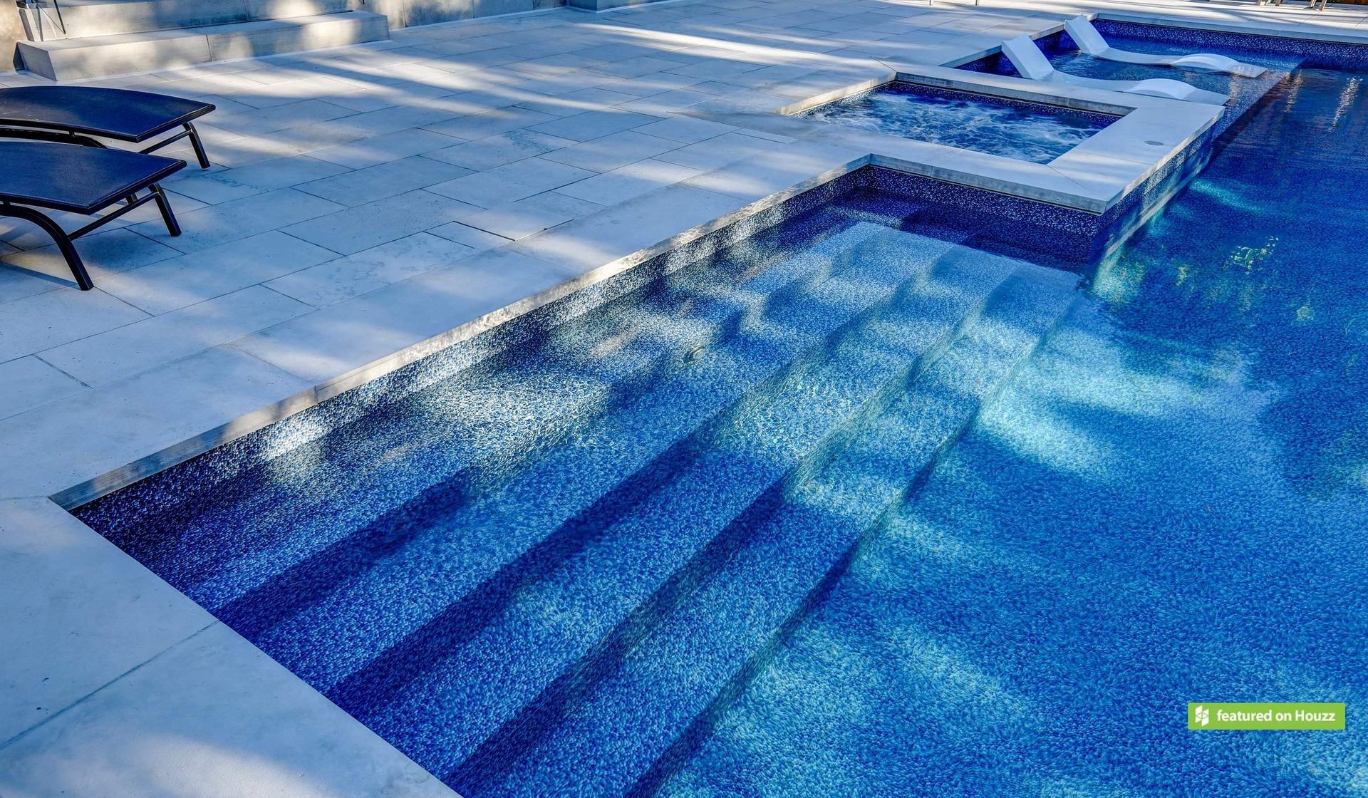 A tiled swimming pool with steps leading into blue water, adjacent to a spa and a paved pool deck with lounge chairs.