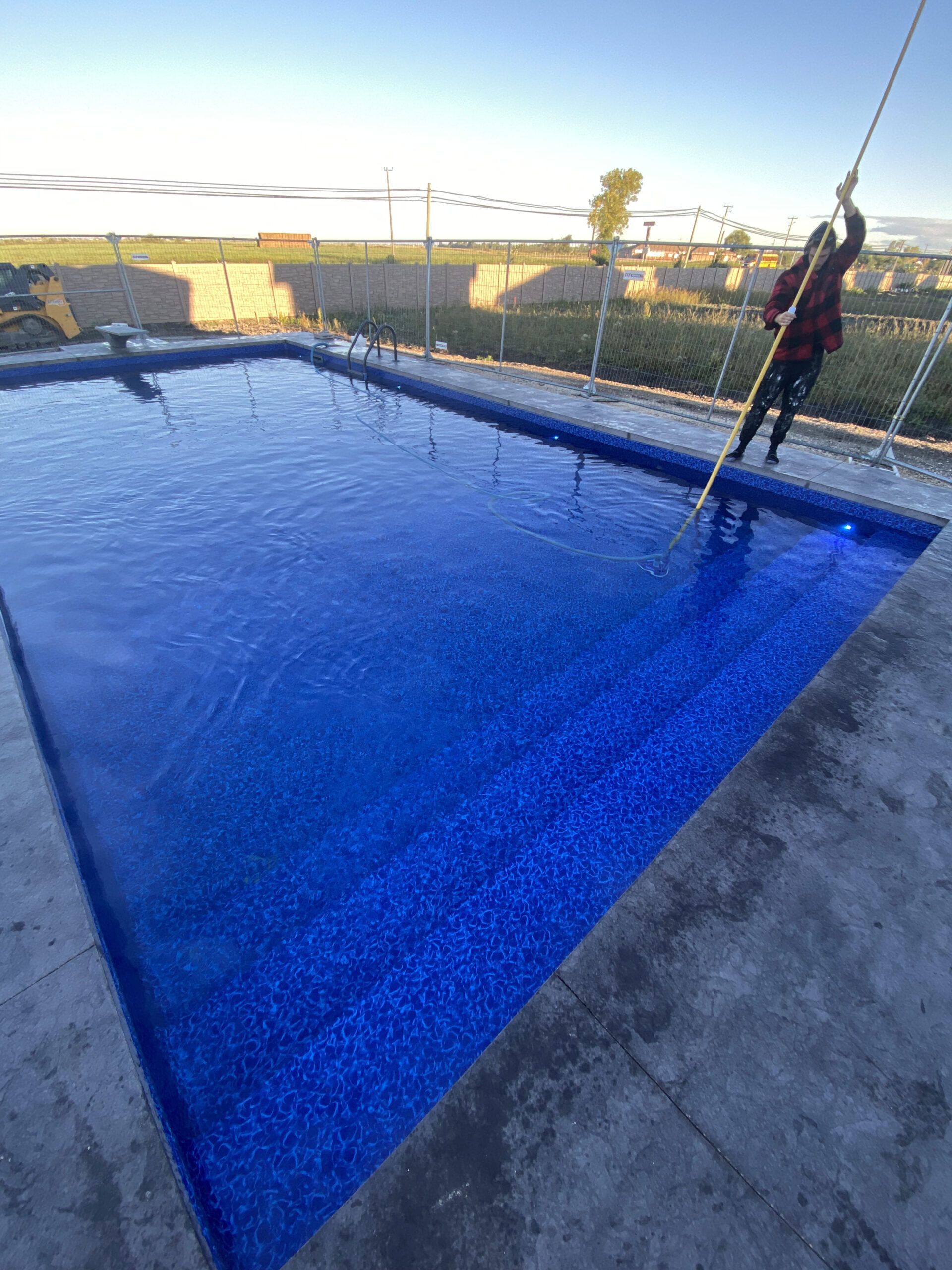 A person stands on the edge of a bright blue tiled swimming pool, using a long pole to clean the water.