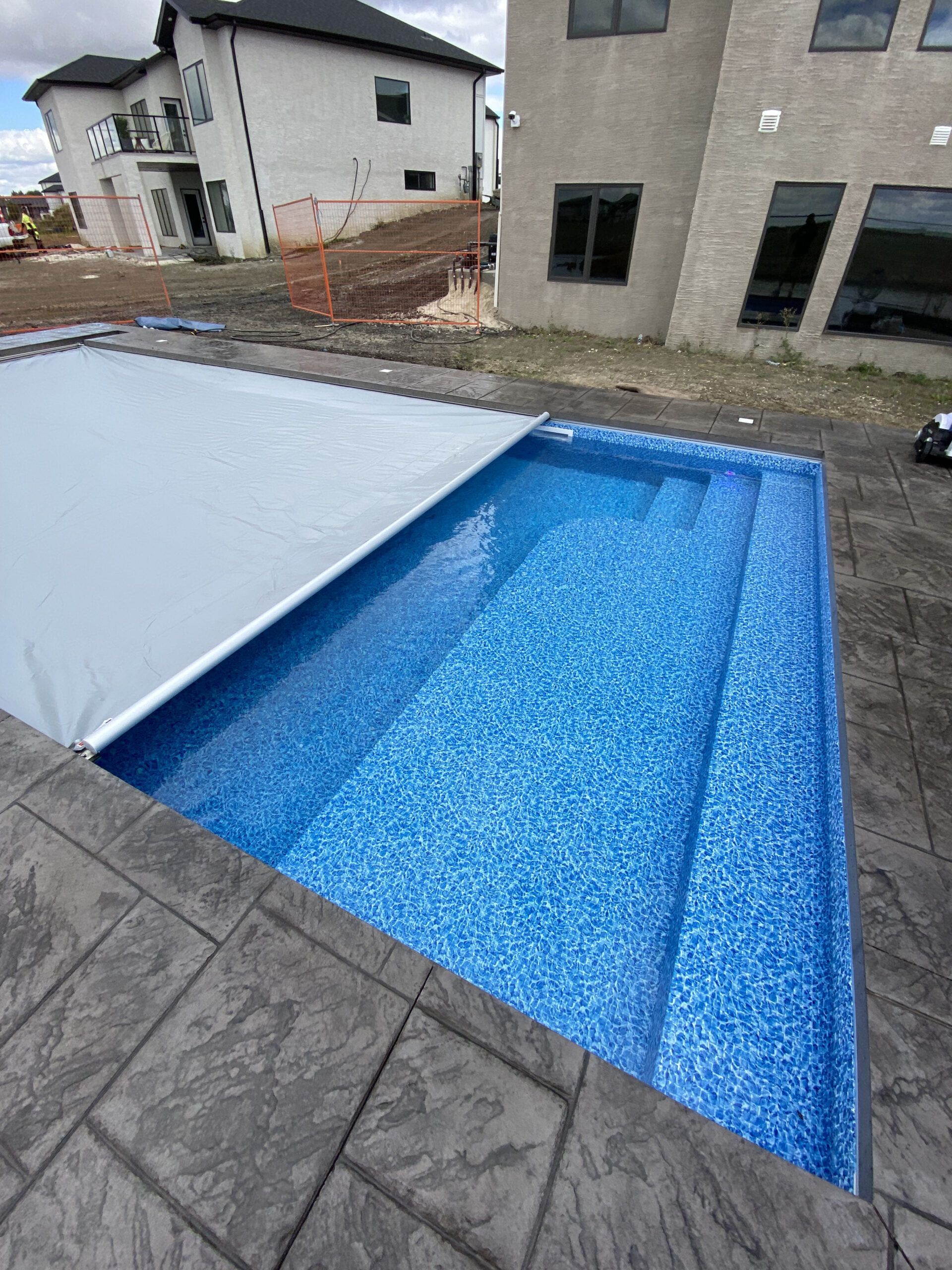 A rectangular swimming pool with a blue tile interior, partially covered by a gray retractable cover on a stone patio.