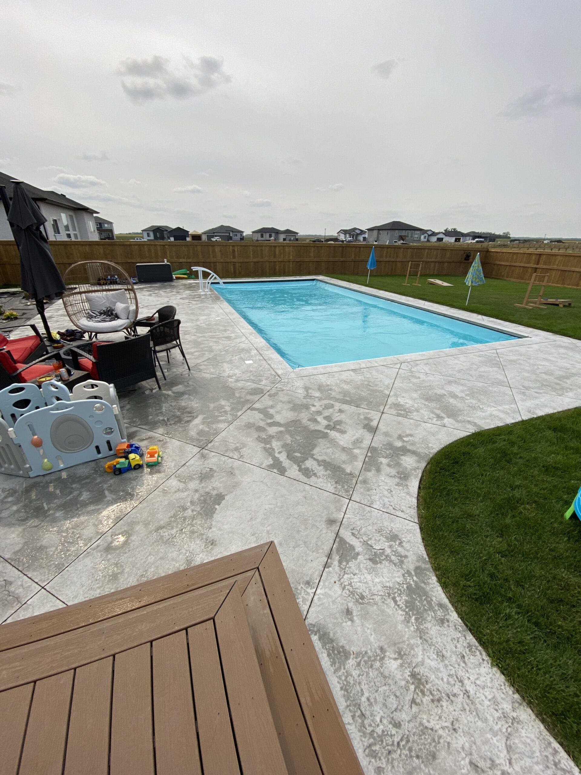 An outdoor backyard with a rectangular swimming pool surrounded by a grey patterned patio, with lounge furniture.