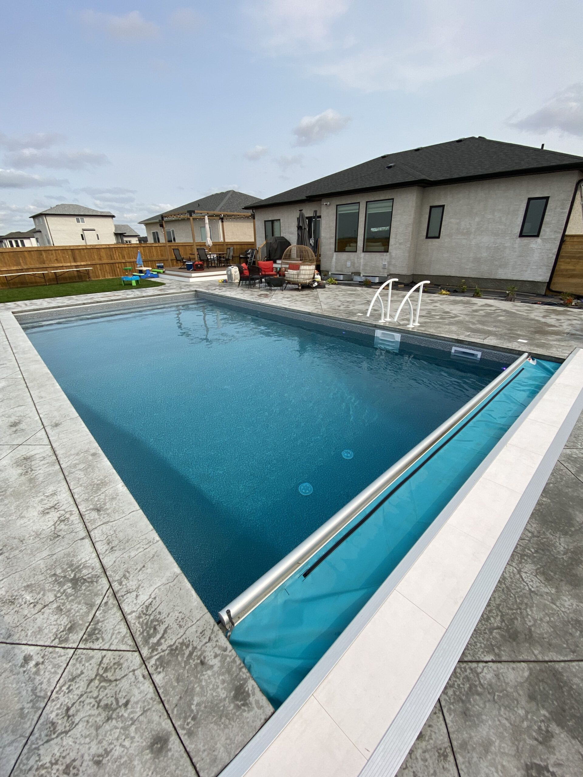 A rectangular backyard swimming pool with a partially rolled-out teal cover, set in a stone patio beside a house.