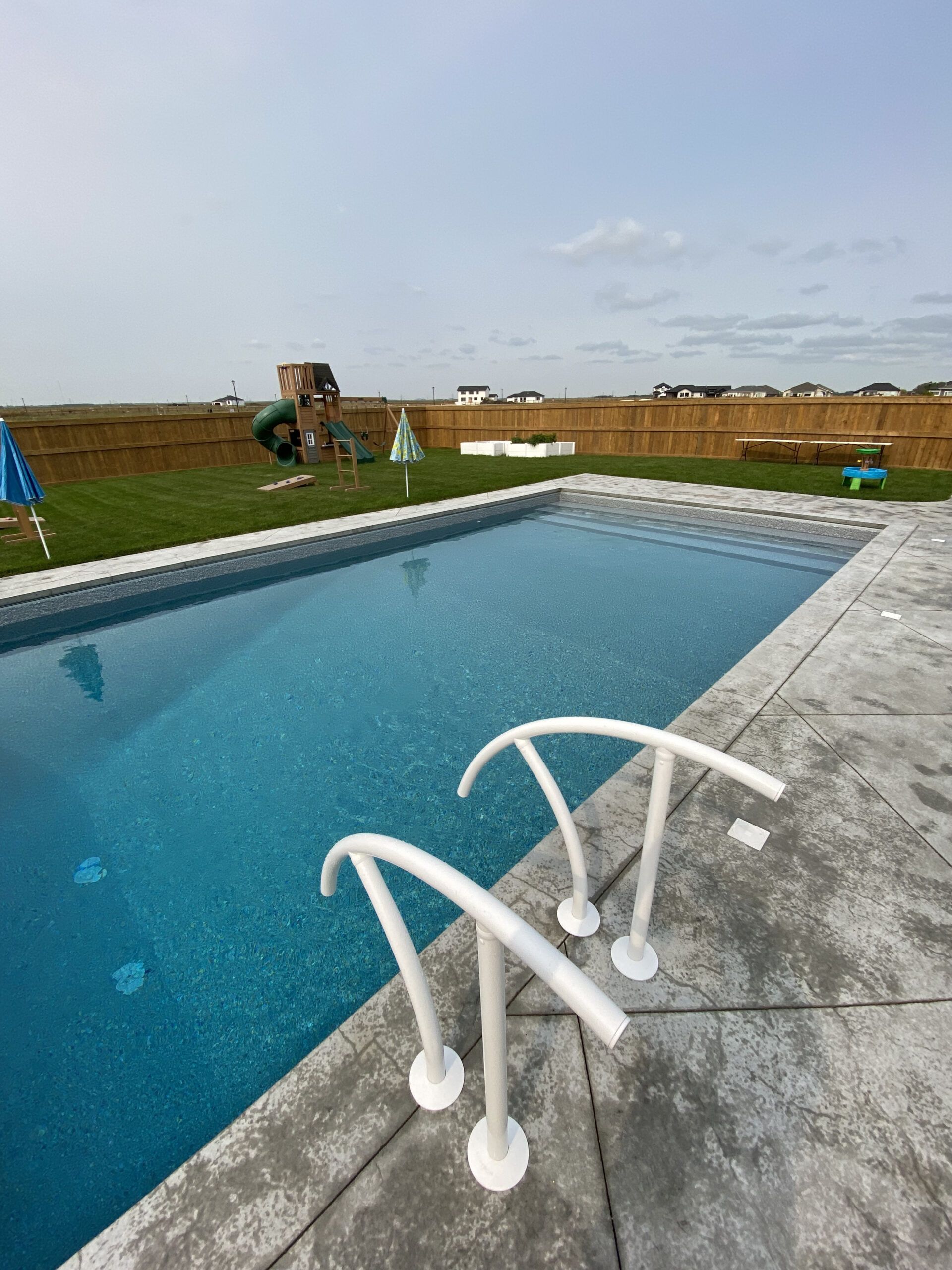 An outdoor swimming pool with light blue water and two white arched railings on a grey concrete deck.