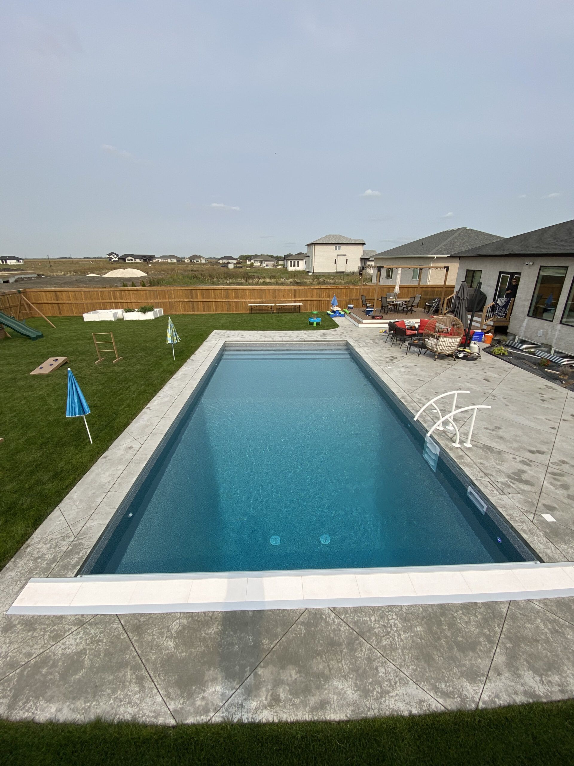 A rectangular backyard swimming pool with blue water and a gray concrete patio, set against a suburban neighborhood.