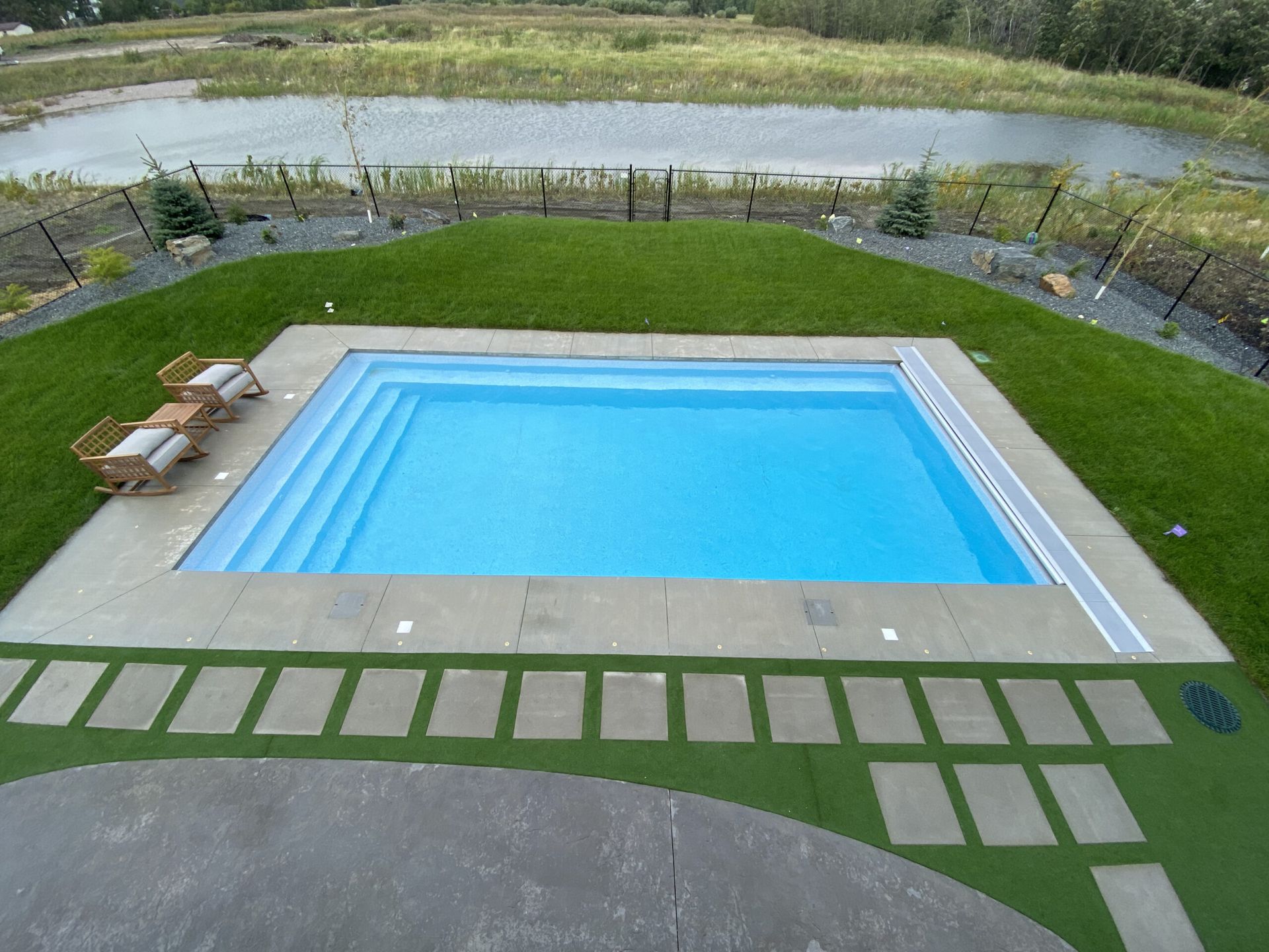 An aerial view of a rectangular, light-blue swimming pool set in a green backyard with a pond in the distance.