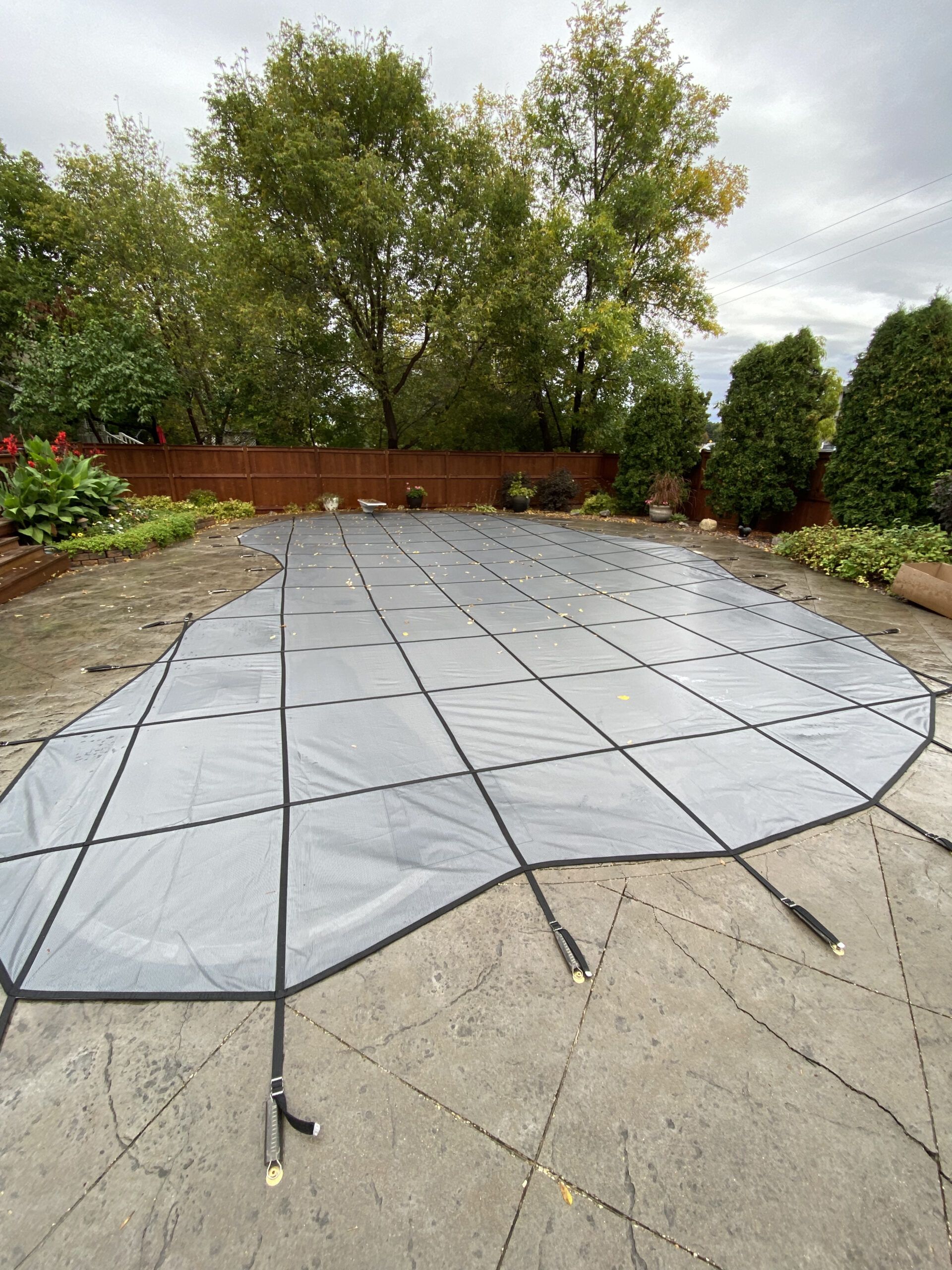 A gray swimming pool safety cover stretched taut over a backyard pool on a patio, surrounded by trees and a wooden fence.