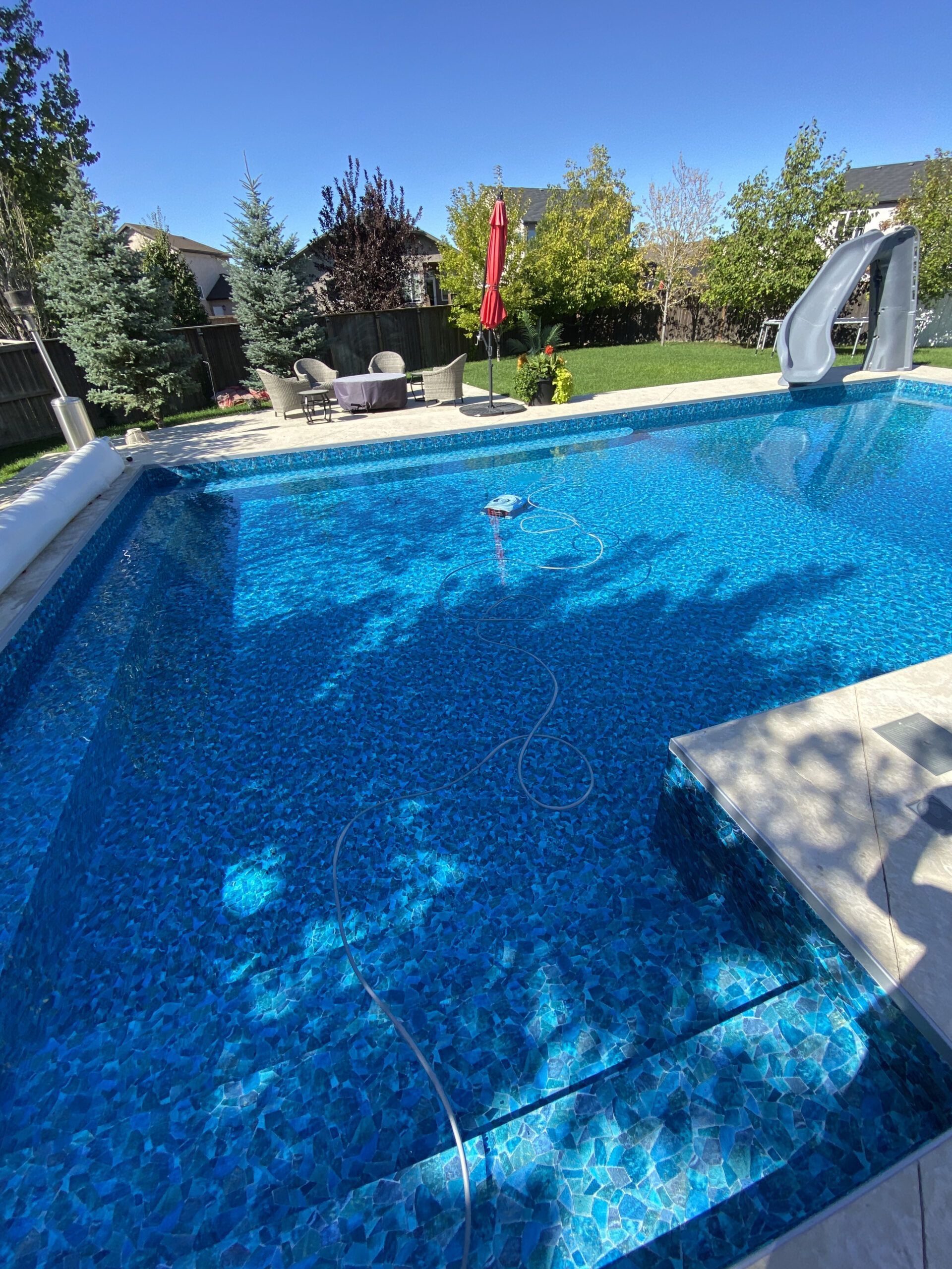 A sunlit backyard swimming pool with a blue tiled liner, a water slide, and a seating area with a fire pit in the back.
