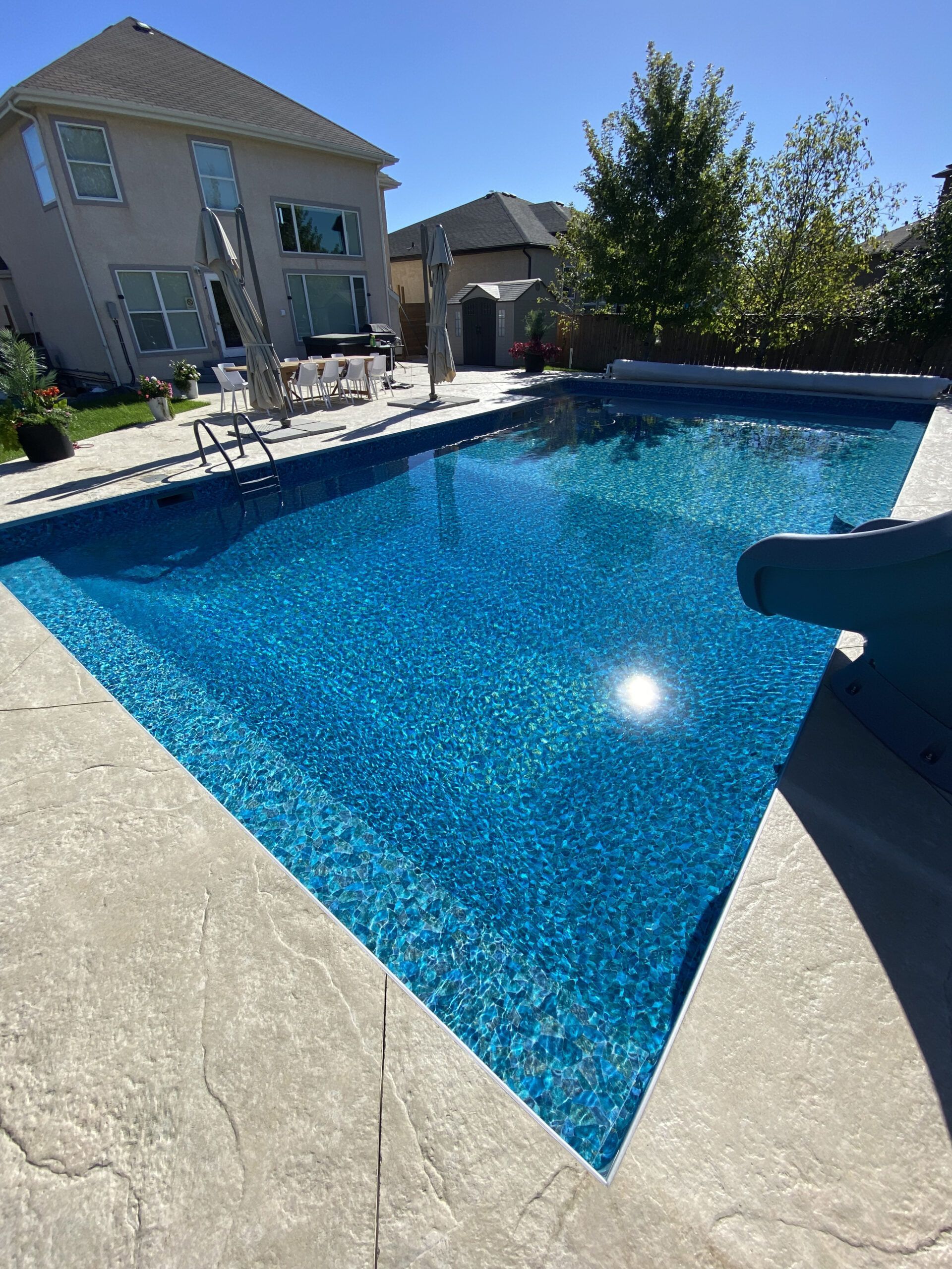 A bright blue backyard swimming pool with a slide, stone patio, and a two-story suburban house in the background.