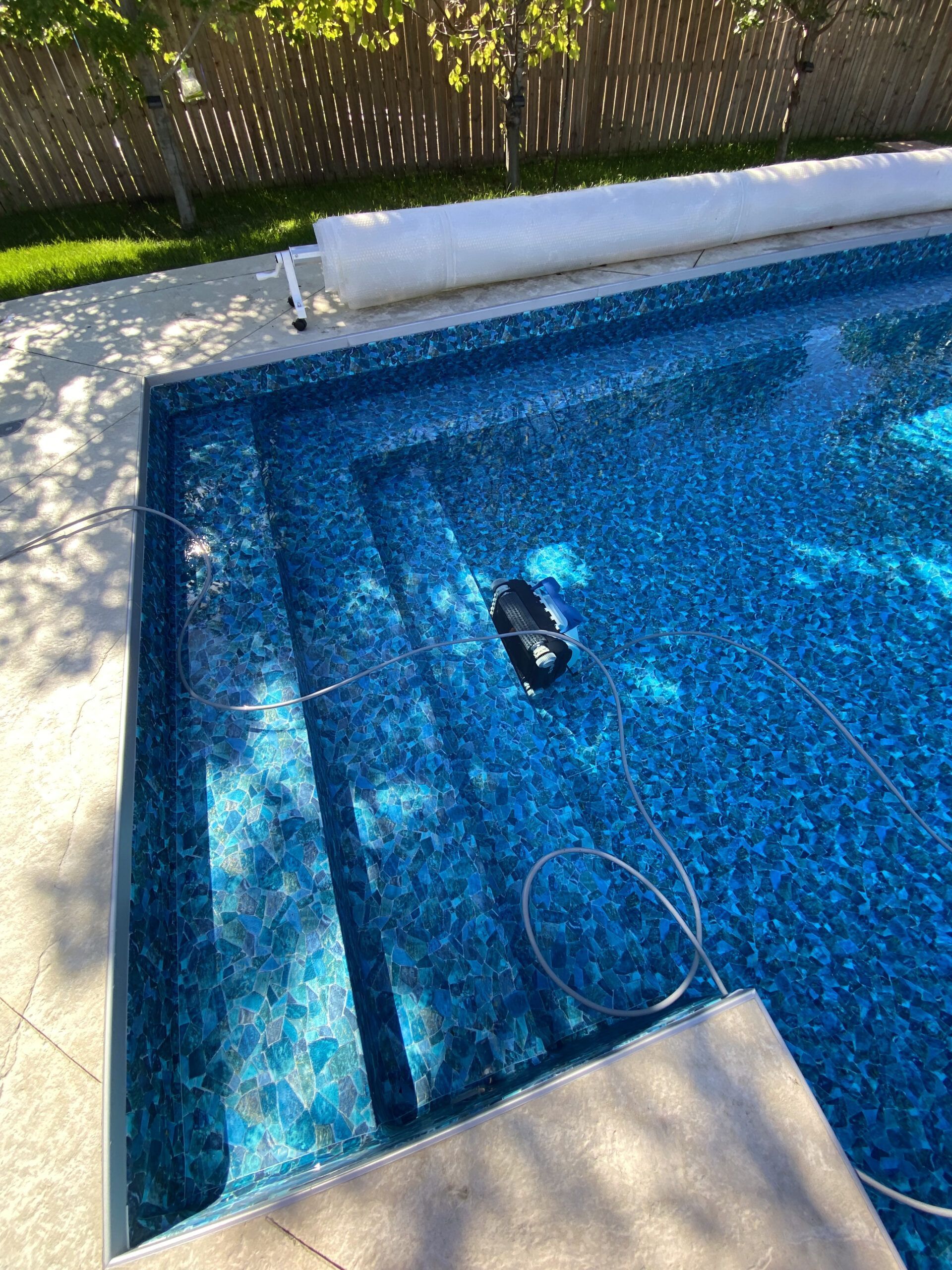An automatic robotic pool cleaner sits on the submerged steps of a blue tiled swimming pool in a sunny backyard.