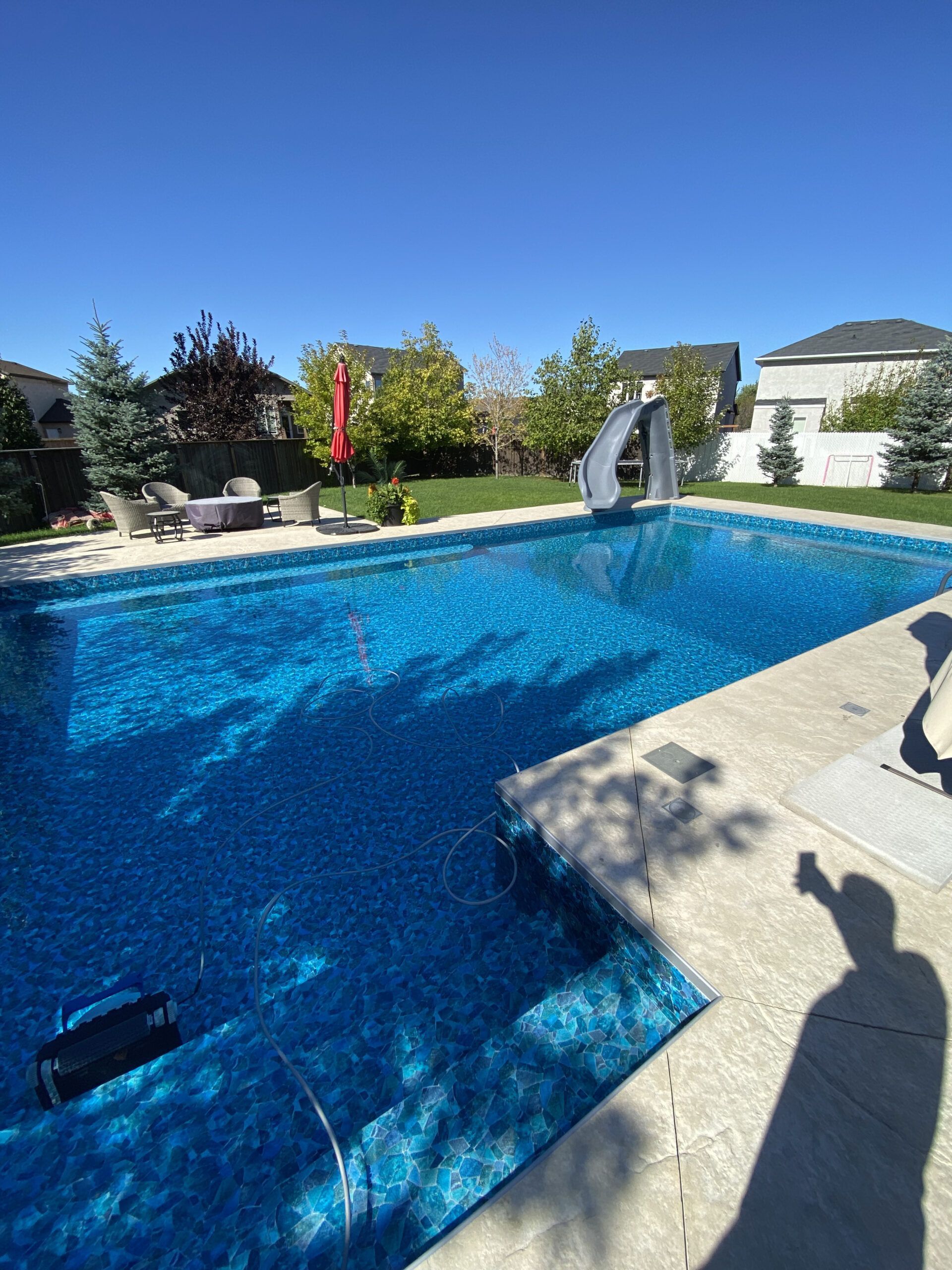 A blue swimming pool with a slide in a sunny backyard setting with patio furniture.