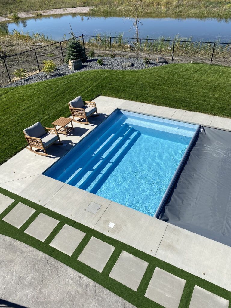 A rectangular swimming pool with blue steps sits in a concrete patio beside two chairs, overlooking a pond.