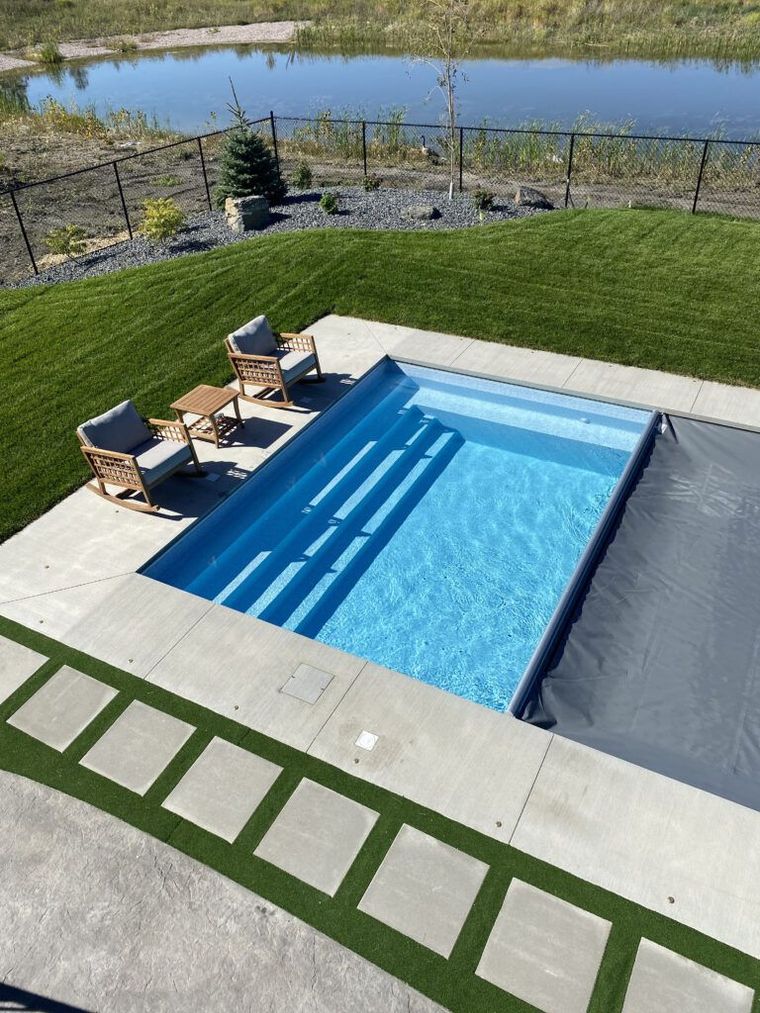 An outdoor rectangular pool with steps and a gray cover, next to patio chairs on a concrete deck beside a grassy yard.