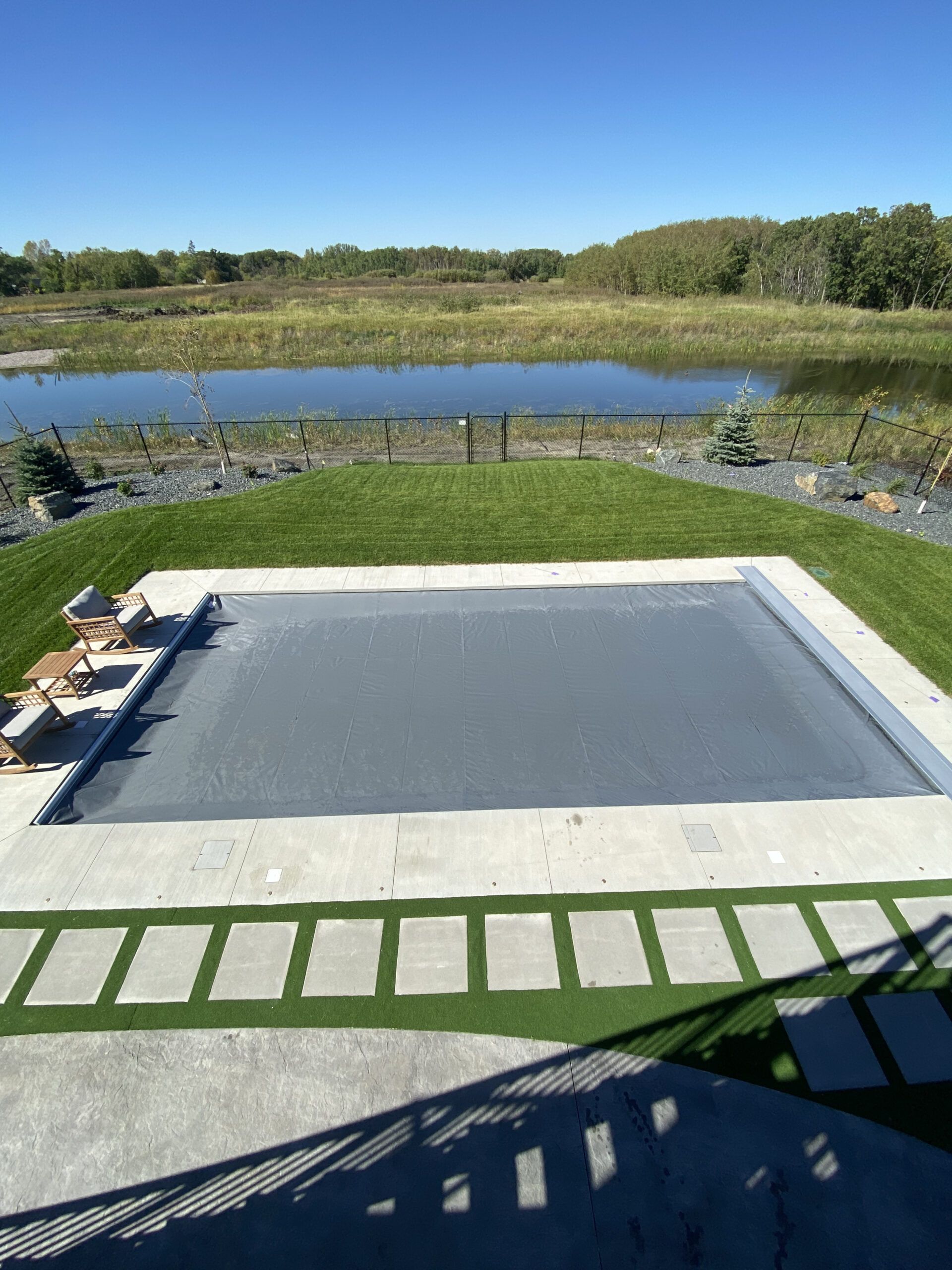 A covered rectangular pool set in a backyard lawn with stone pavers and a pond in the background under a blue sky.