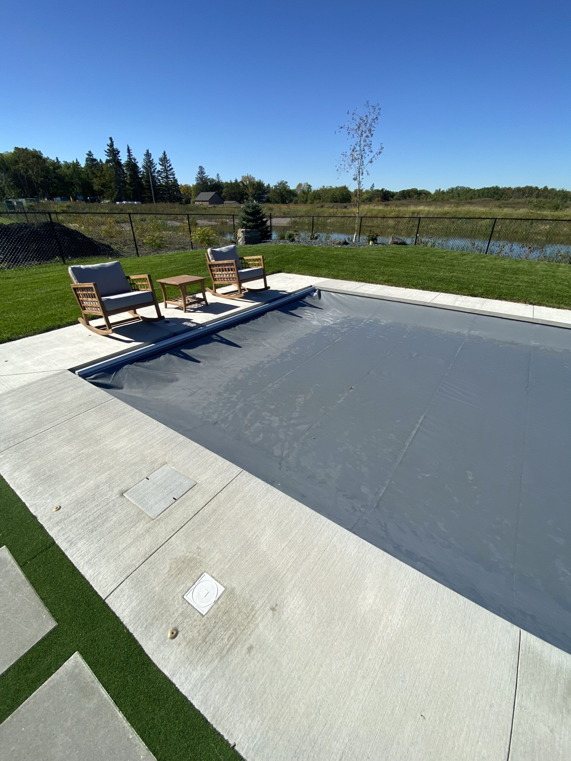 A gray pool cover spans a rectangle pool on a patio with two chairs, overlooking a grassy field and distant trees.