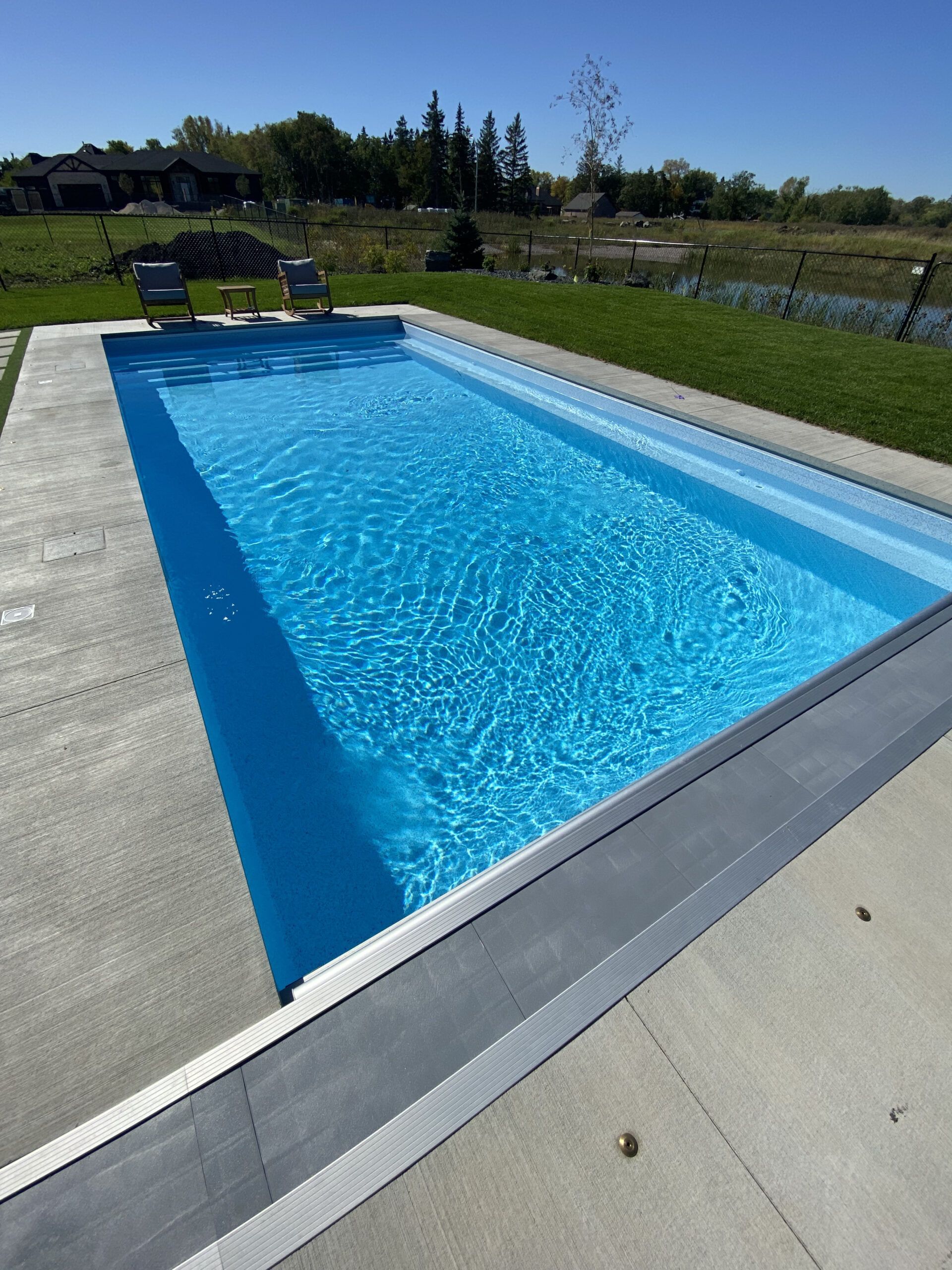 A bright blue rectangular swimming pool with a concrete deck, two chairs at the far end, and a grassy yard beyond.