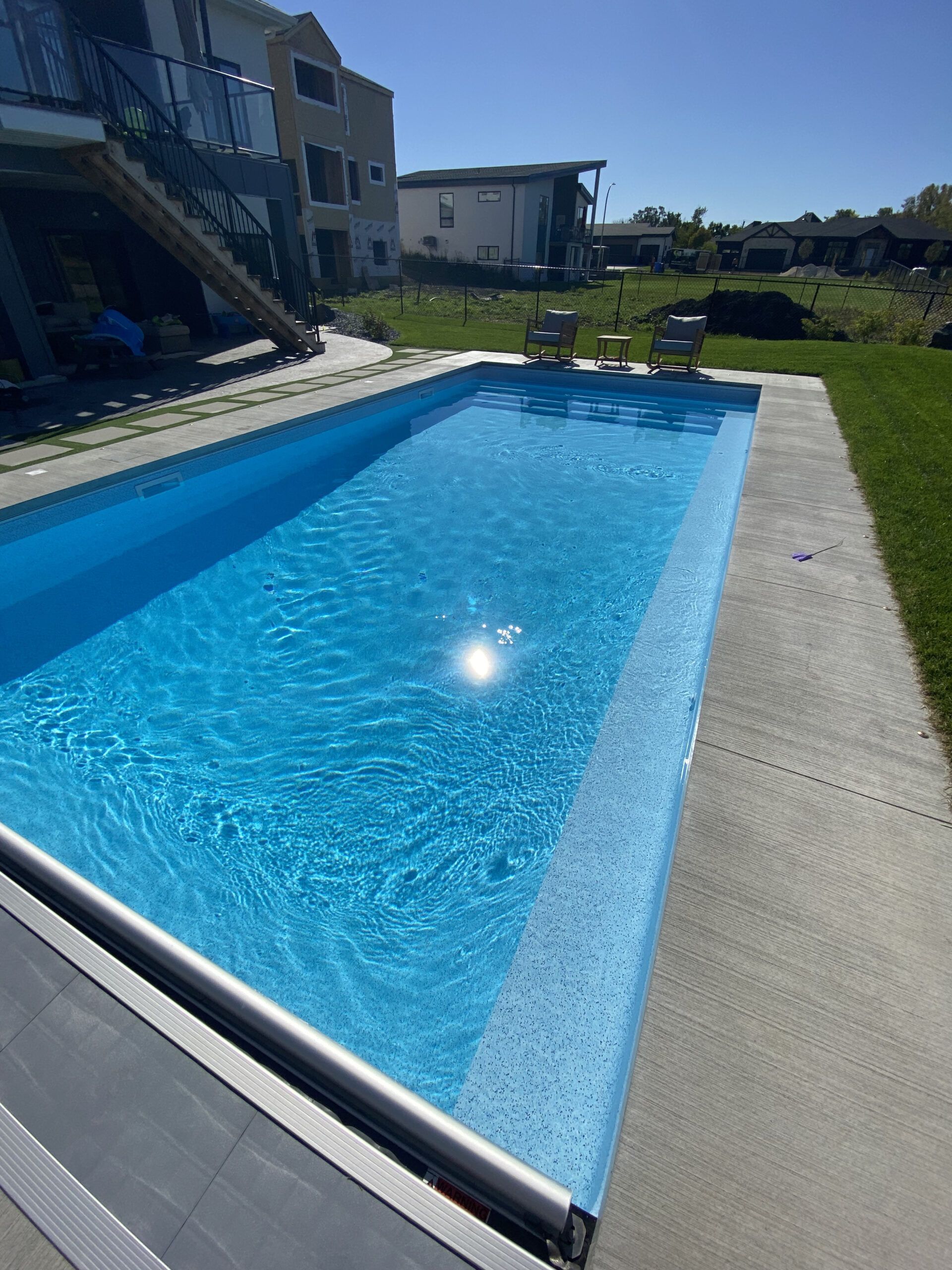 A bright blue rectangular swimming pool in a residential backyard with a concrete deck and a partial pool cover.