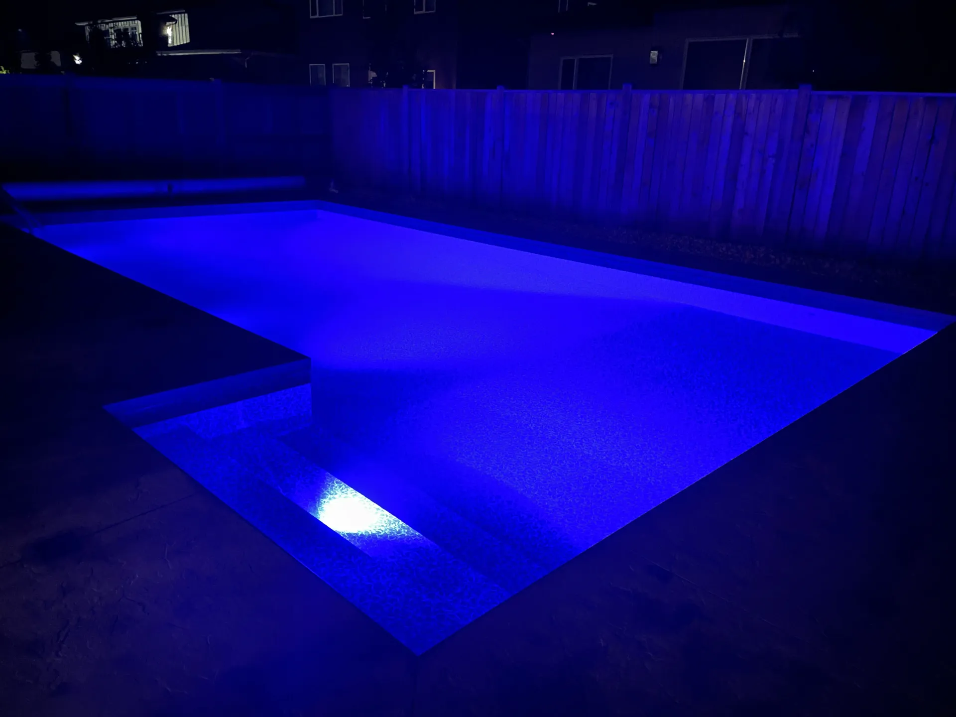 A rectangular swimming pool illuminated by vibrant blue LED lights at night, viewed from a patio next to a wooden fence.