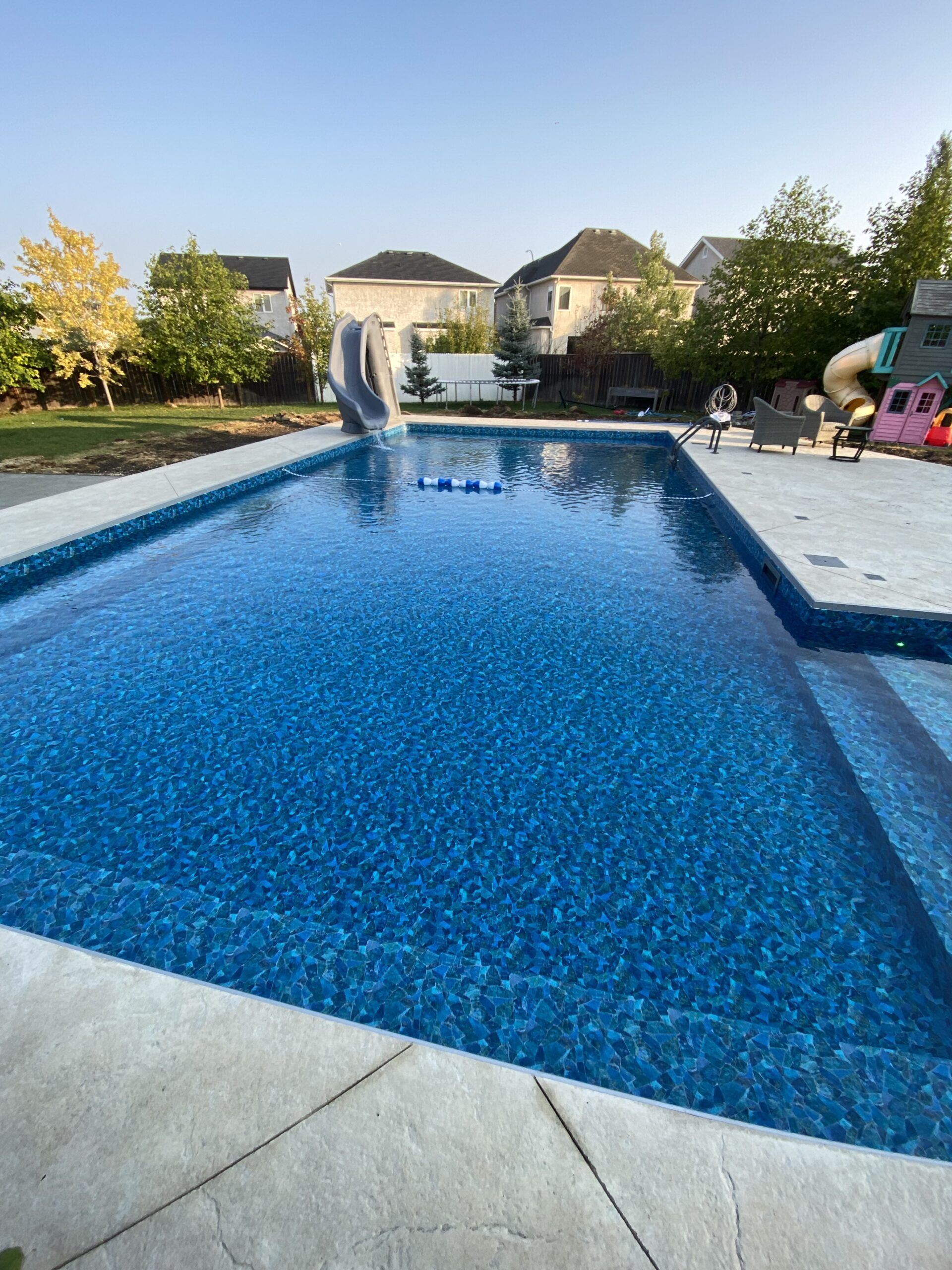 A rectangular inground pool with dark blue patterned tiles, surrounded by a light-colored concrete deck on a sunny day.