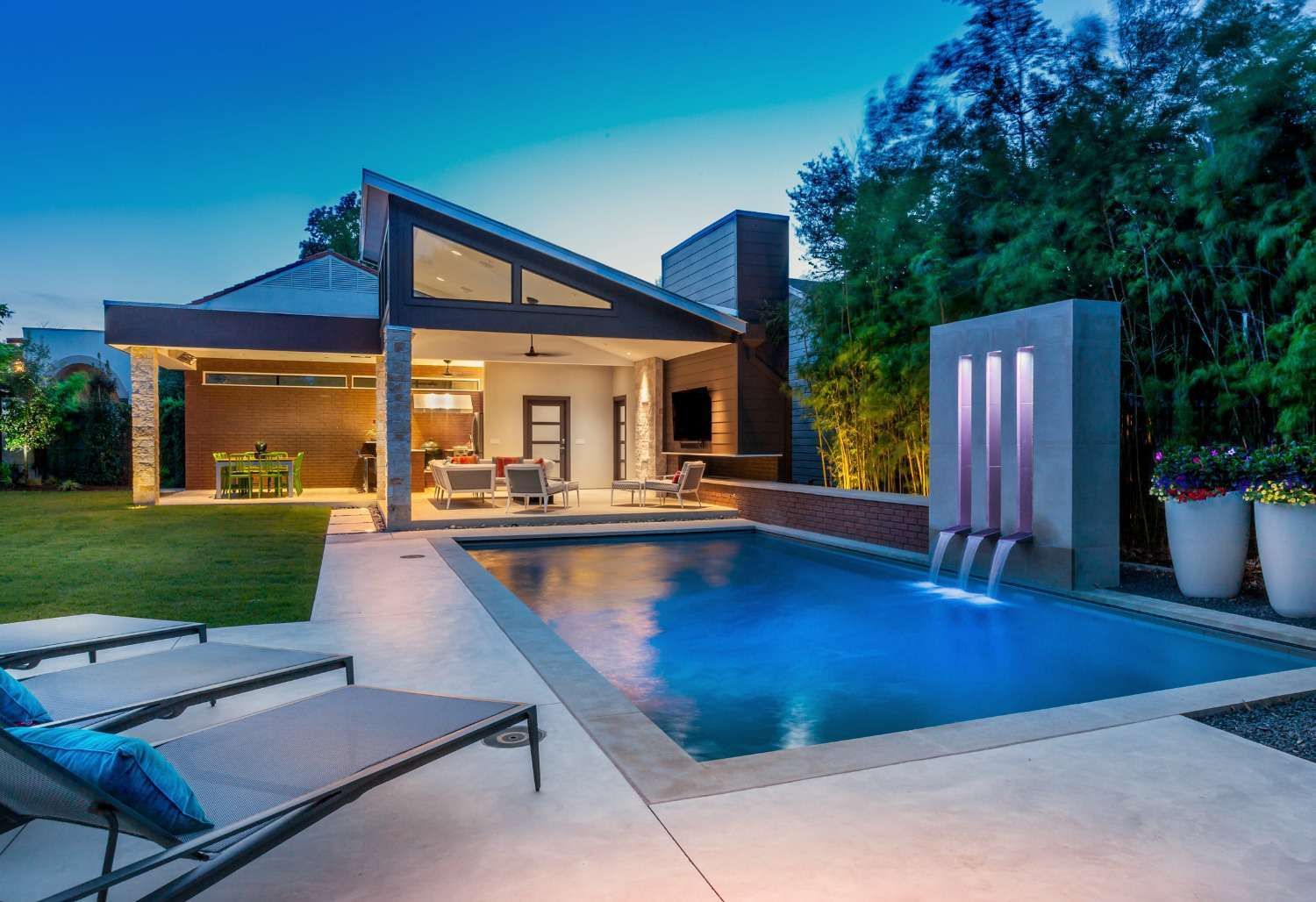 A backyard patio with a pool, a modern open-air living space, and a lighted water feature at dusk.