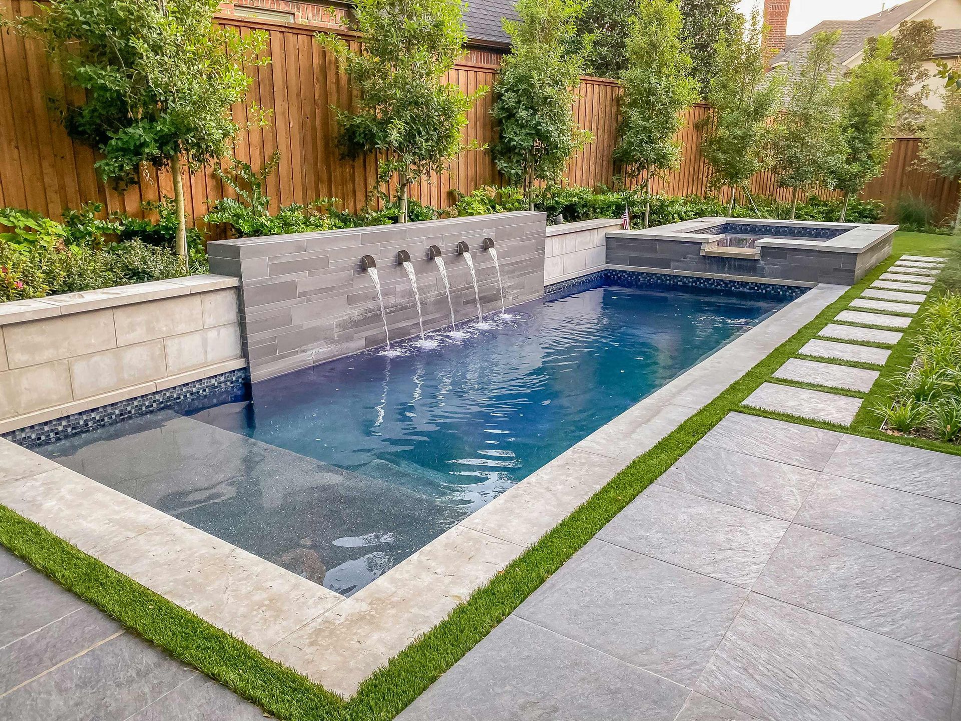 A rectangular swimming pool with a stone wall water feature, travertine coping, and a path of stepping stones on the grass.