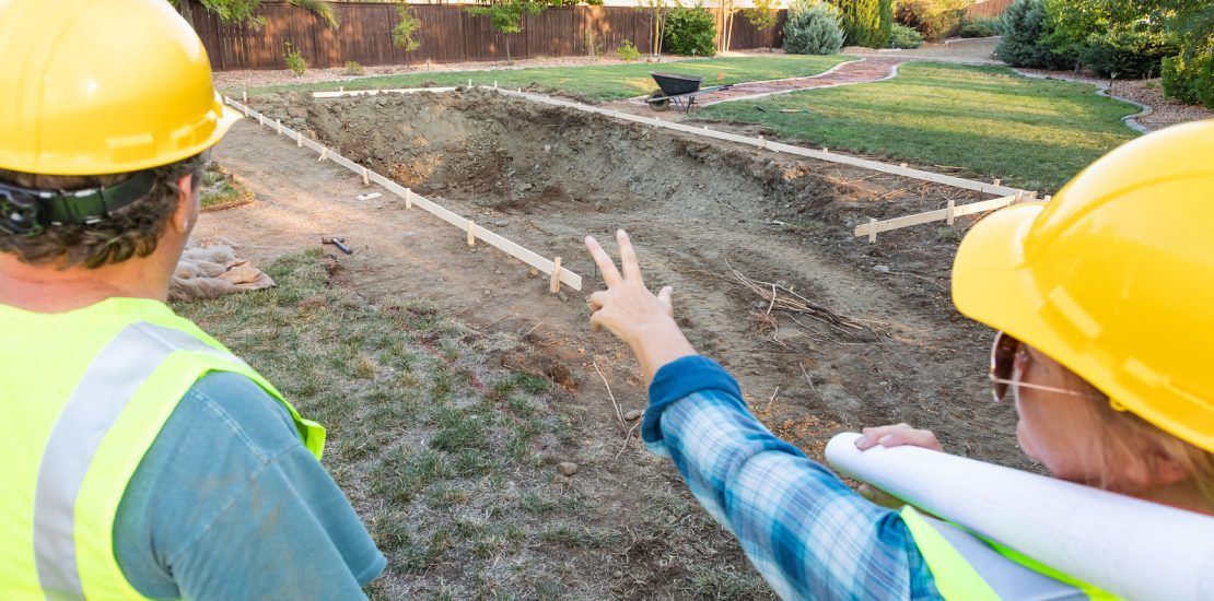 Two people in hard hats and reflective vests look at a trench marked with stakes in a residential yard.