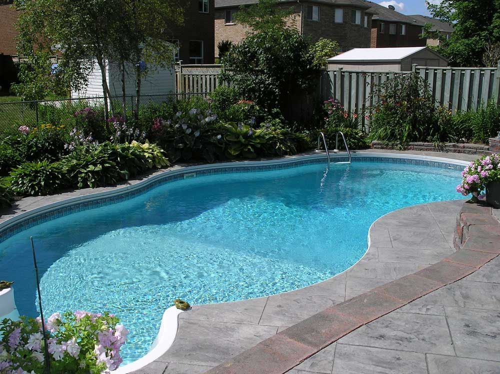 A backyard swimming pool with a curved edge, blue water, and a stamped concrete patio surrounded by lush green garden beds.
