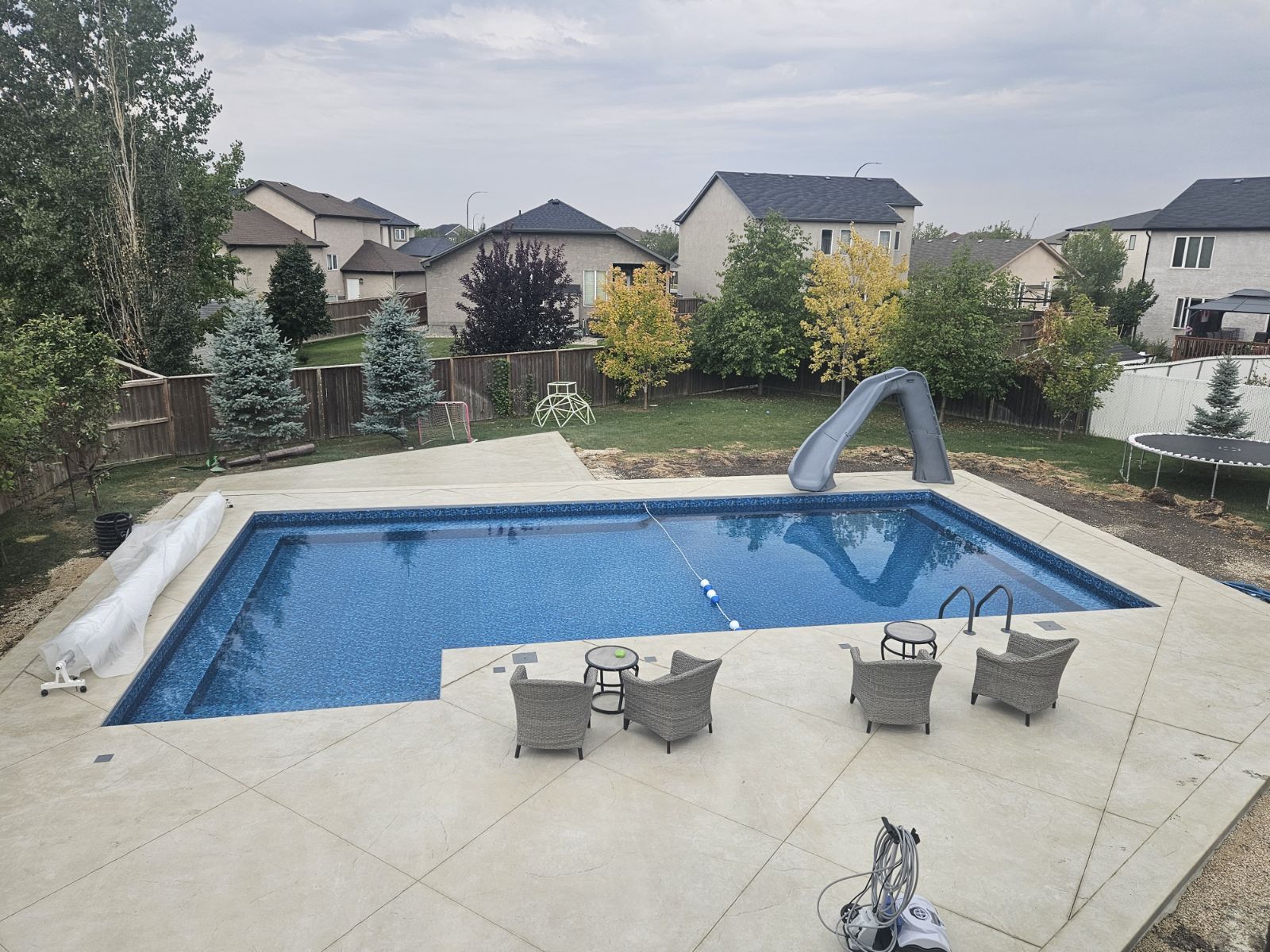 An L-shaped swimming pool with a slide, surrounded by a light-colored concrete patio with furniture in a backyard.