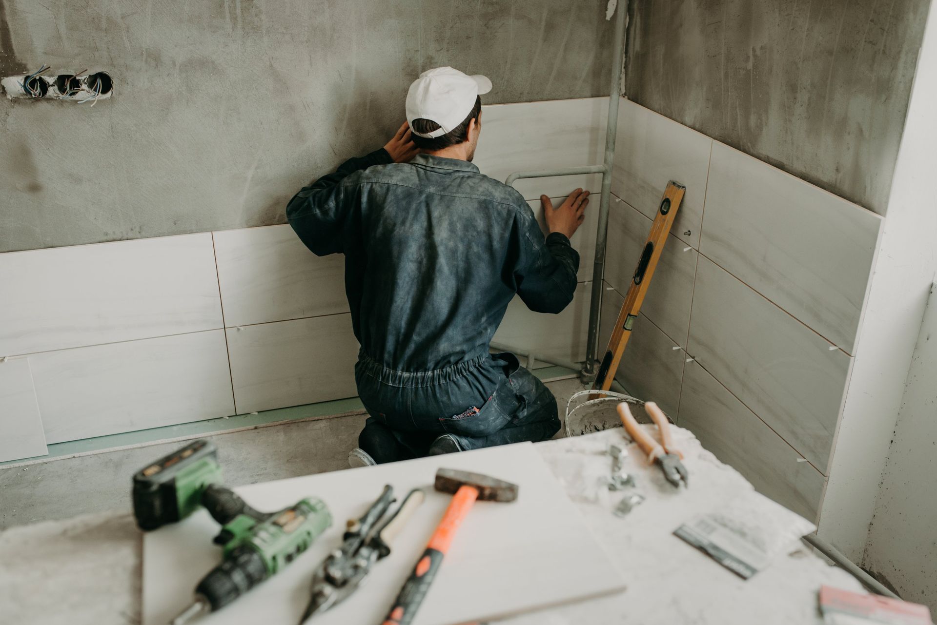 A worker in dark coveralls and a white cap installs light-colored wall panels in a room with tools on a table nearby.