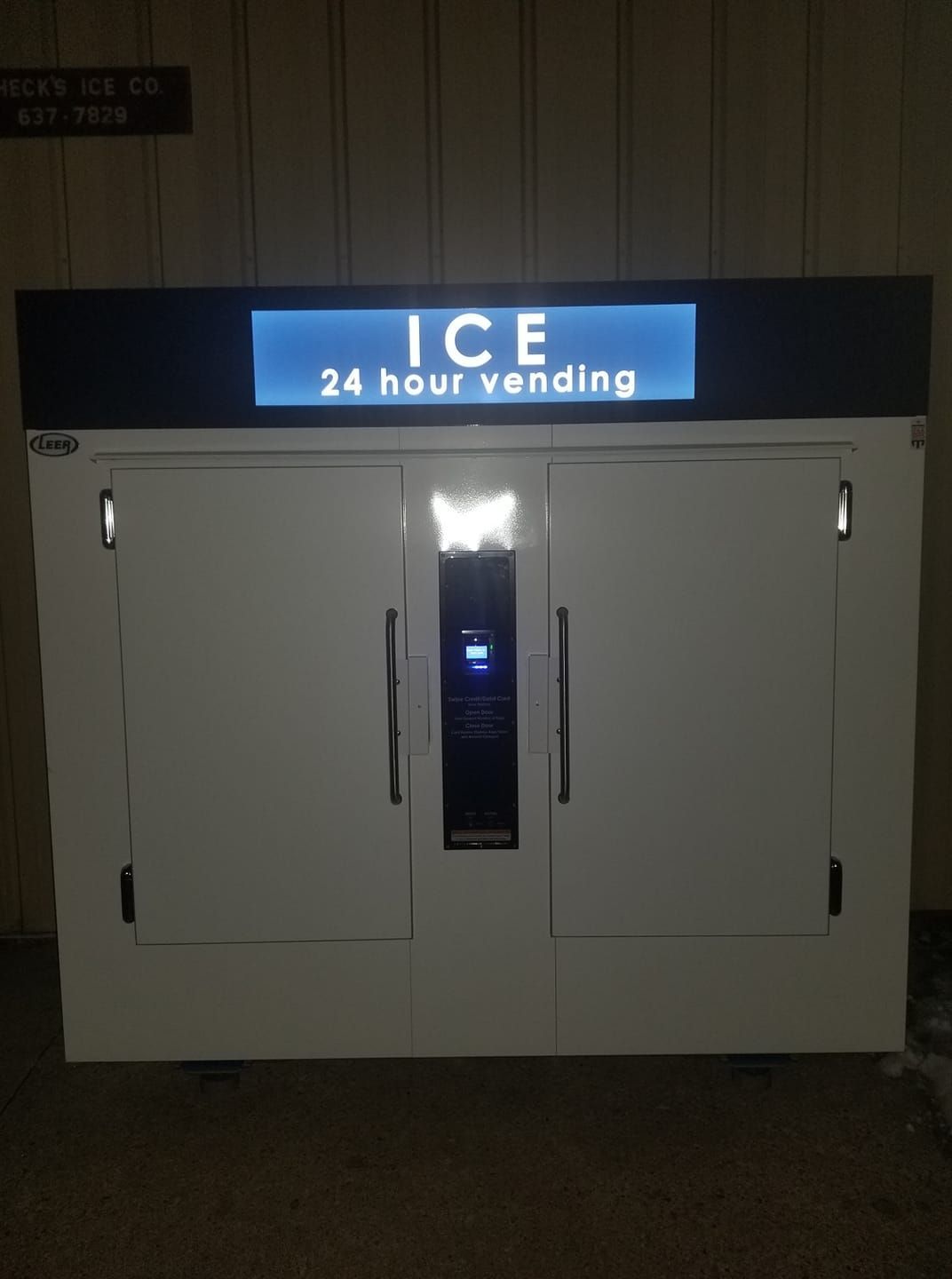 White ice vending machine with a lit sign above that reads “ICE 24 HOUR VENDING.”