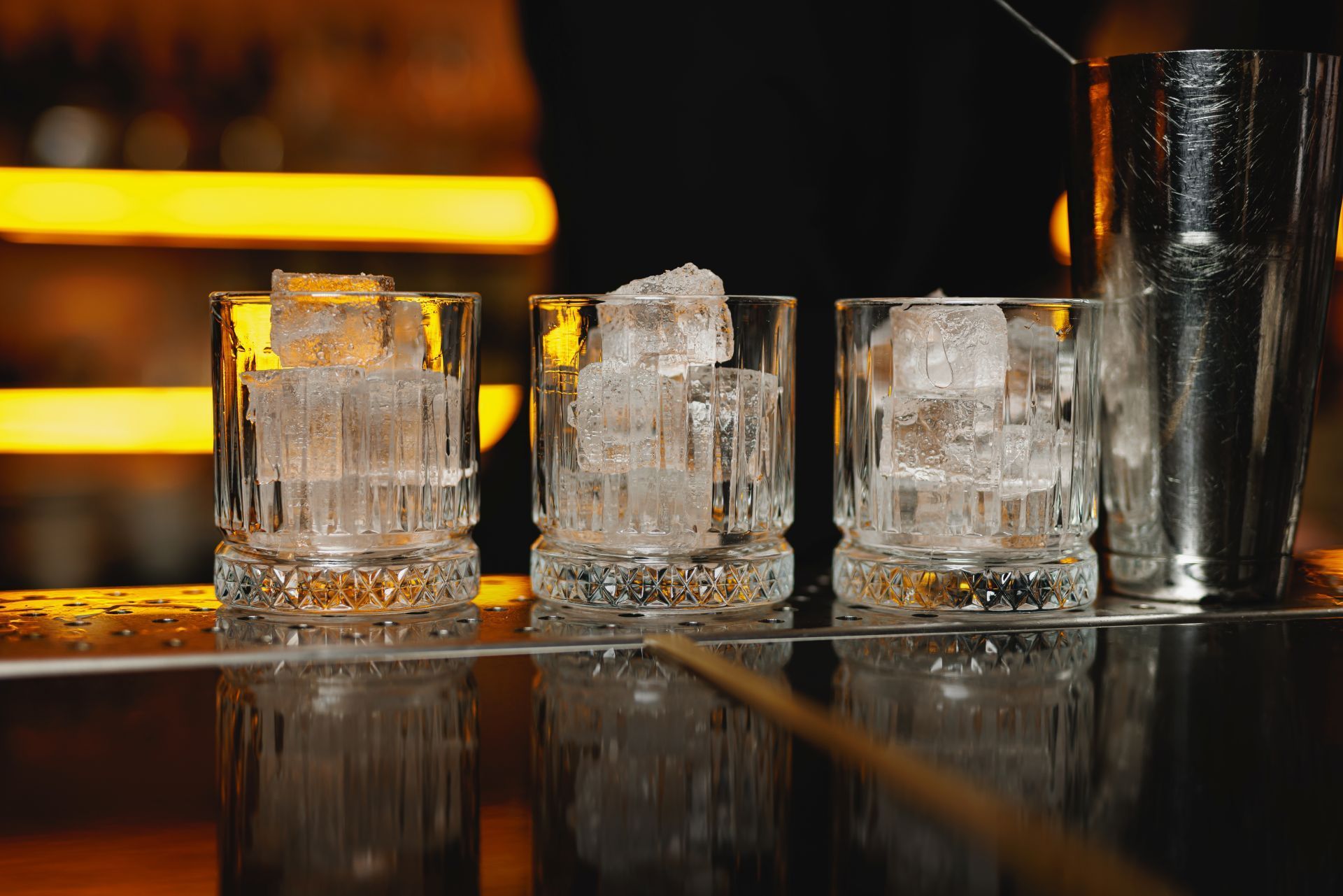 Three rocks glasses with ice cubes on a bar. A metal cocktail shaker is next to the glasses.