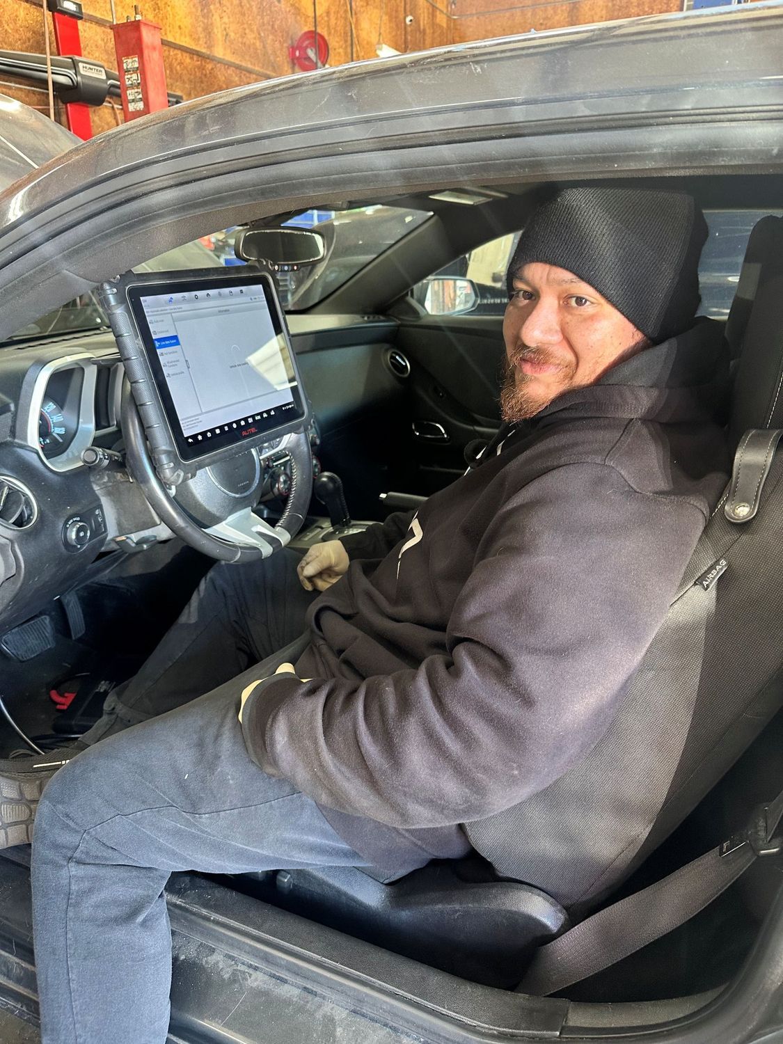 A person wearing a beanie and hoodie sits in a car, using a tablet mounted to the steering wheel in a garage.