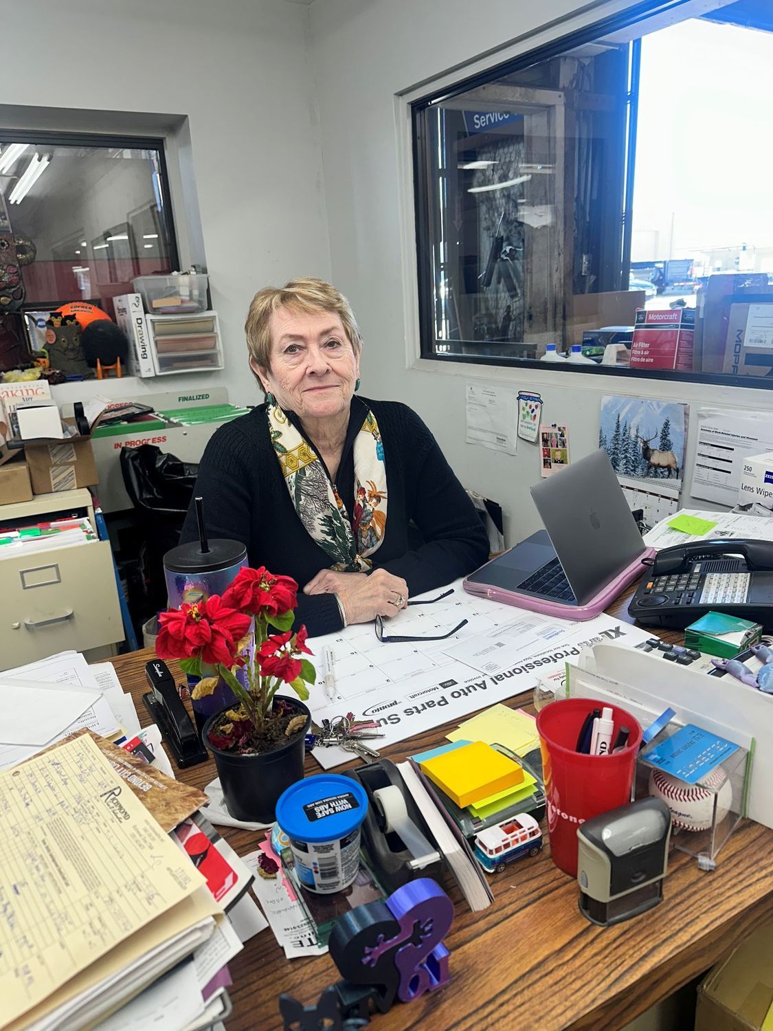A person sits at a cluttered office desk with a laptop, potted plant, and various stationery items in a bright room.