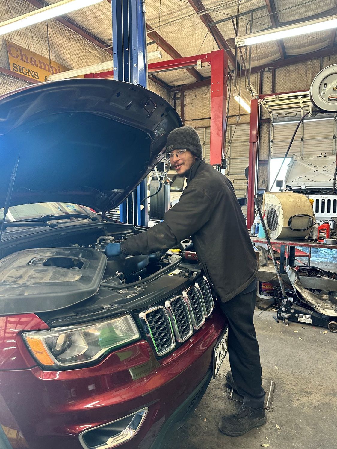 A person wearing a beanie and dark jacket works under the hood of a red Jeep inside an automotive repair shop.