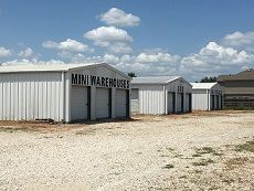 Rows of white, metal mini-warehouse storage units on a gravel lot under a blue sky with scattered clouds.