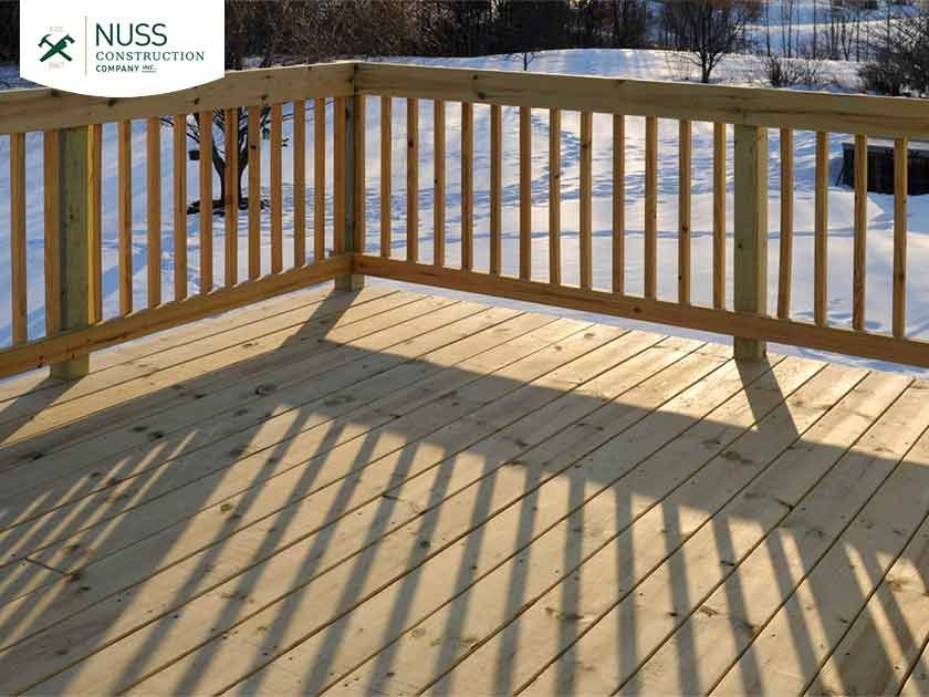 Wooden deck with railings casting long shadows over light-colored planks in a snowy outdoor setting.
