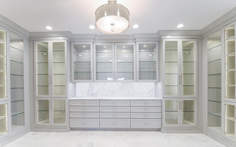 A symmetrical white luxury walk-in closet with glass-front cabinets, internal lighting, marble backsplash, and drawers.