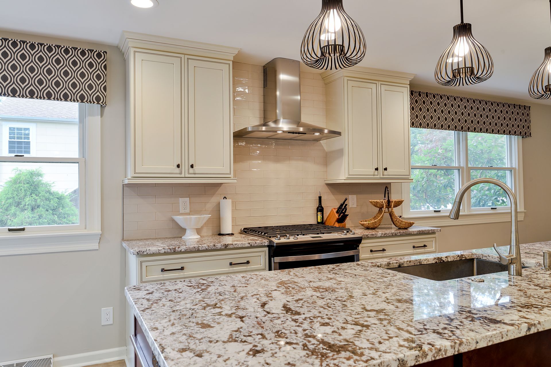 A bright kitchen with white cabinets, speckled granite countertops, a stainless steel hood, and patterned window valances.