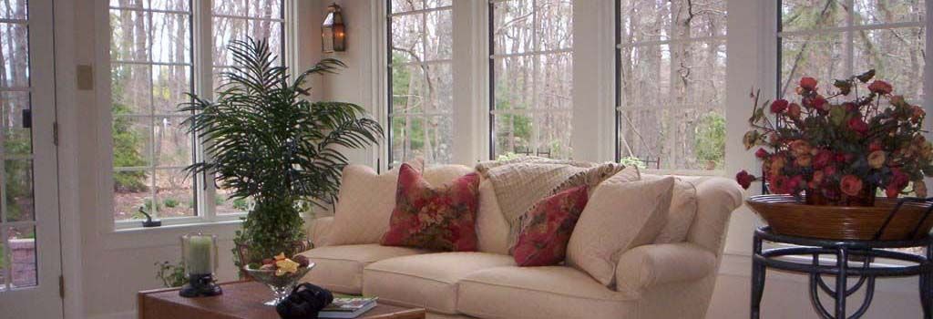 A light-colored sofa with red pillows in a sunroom, featuring a coffee table, a green plant, and a potted floral arrangement.