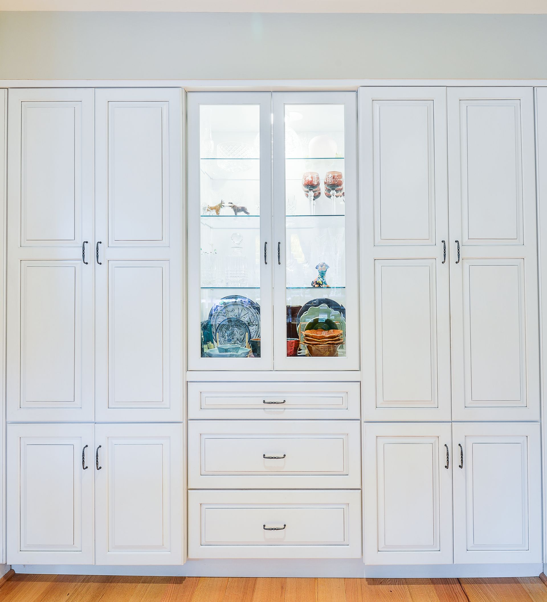 A large white wall unit with glass-front display cabinets, storage doors, and drawers, set on a hardwood floor.
