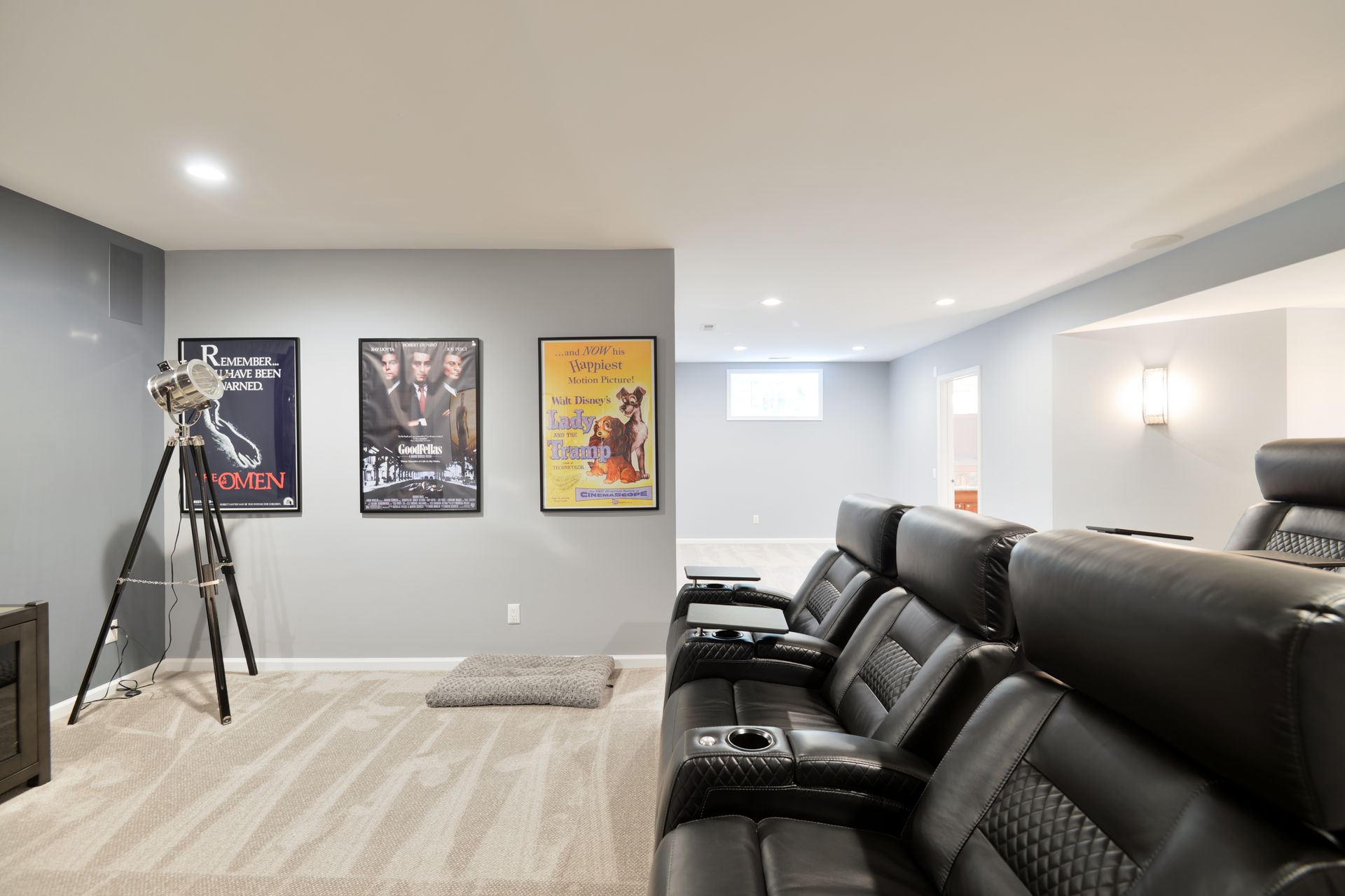 A home theater room with black leather recliners, a tripod spotlight, and three framed movie posters on a gray wall.