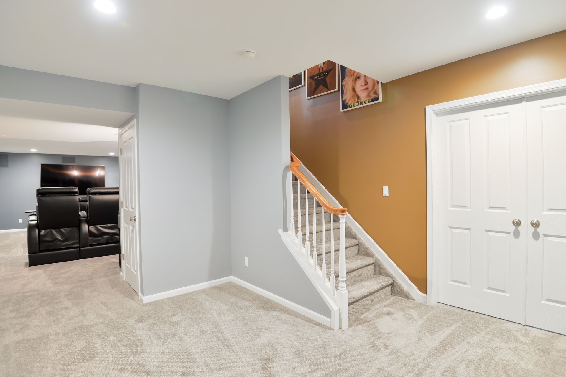 A carpeted basement with gray walls, a staircase with white railings, a tan feature wall, and white double closet doors.