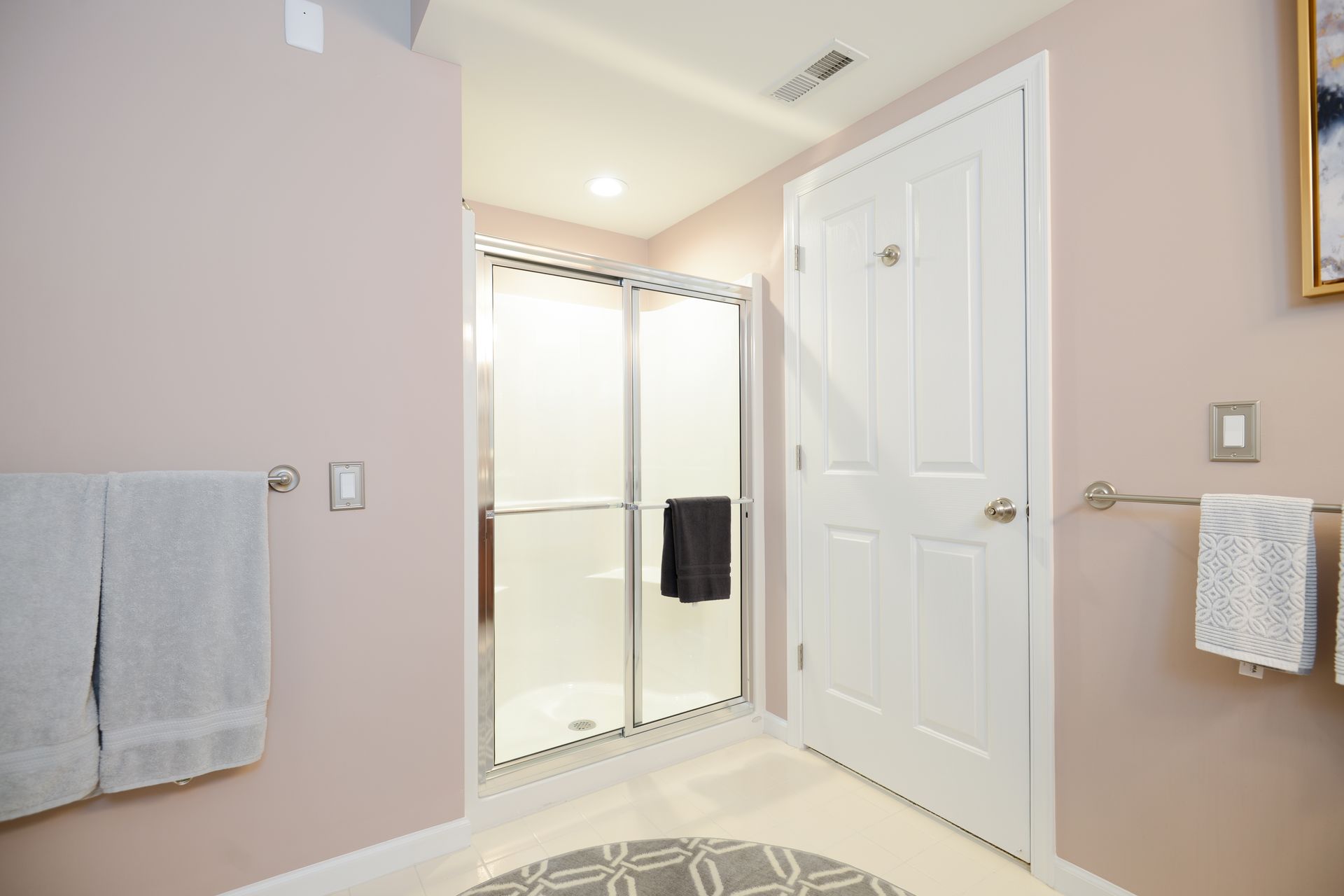A light-pink bathroom featuring a walk-in shower with sliding glass doors, a white door, and hanging towels.