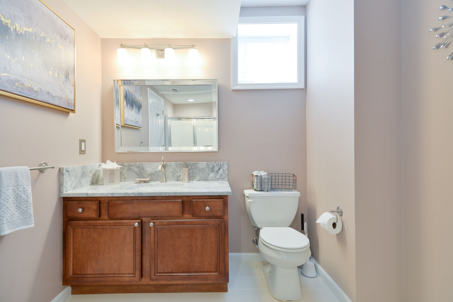 A bathroom with a wooden vanity, a marble-topped counter, a mirror, a toilet, and light pink walls under a small window.