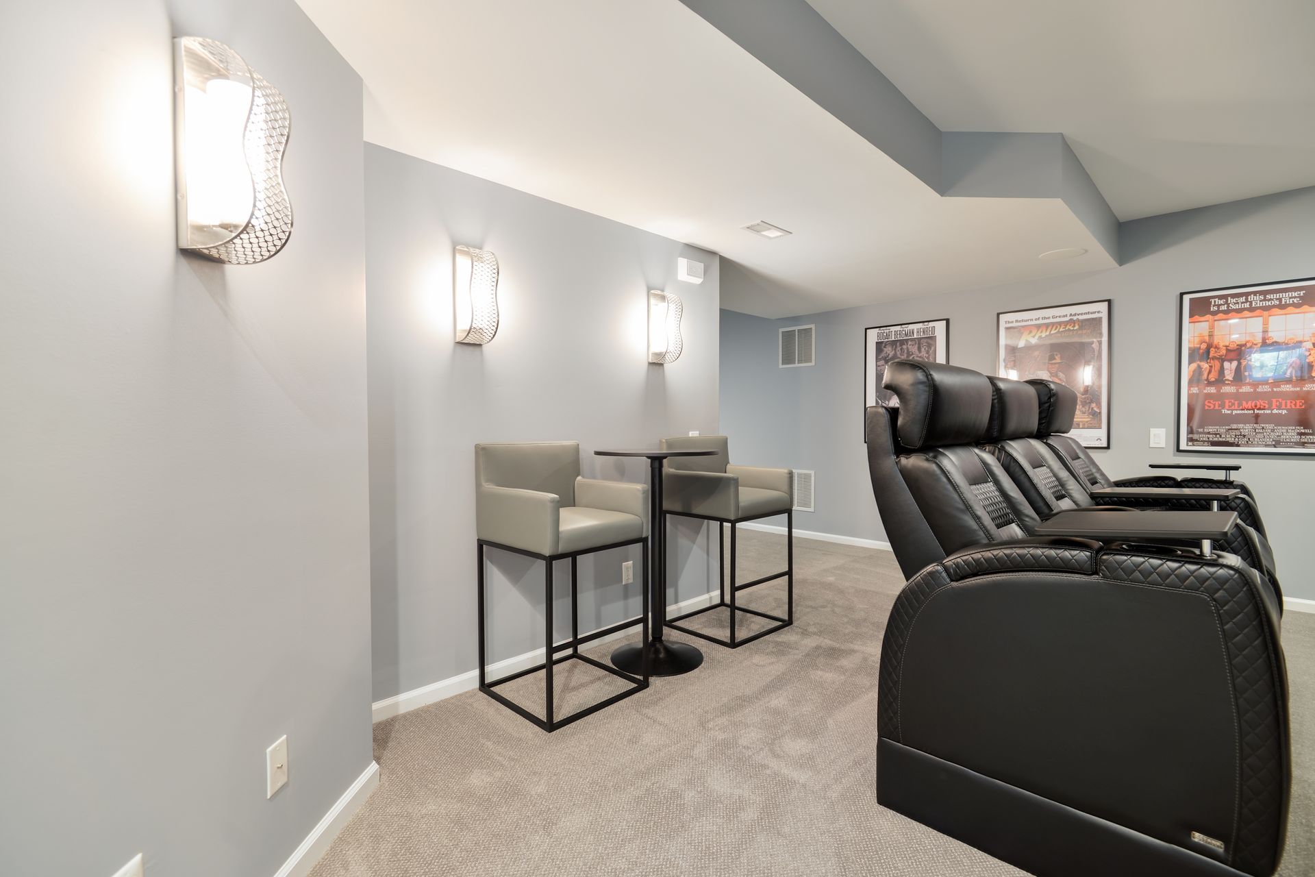 Home theater with black leather seats, a small table with two gray bar stools, and wall sconces on light gray walls.