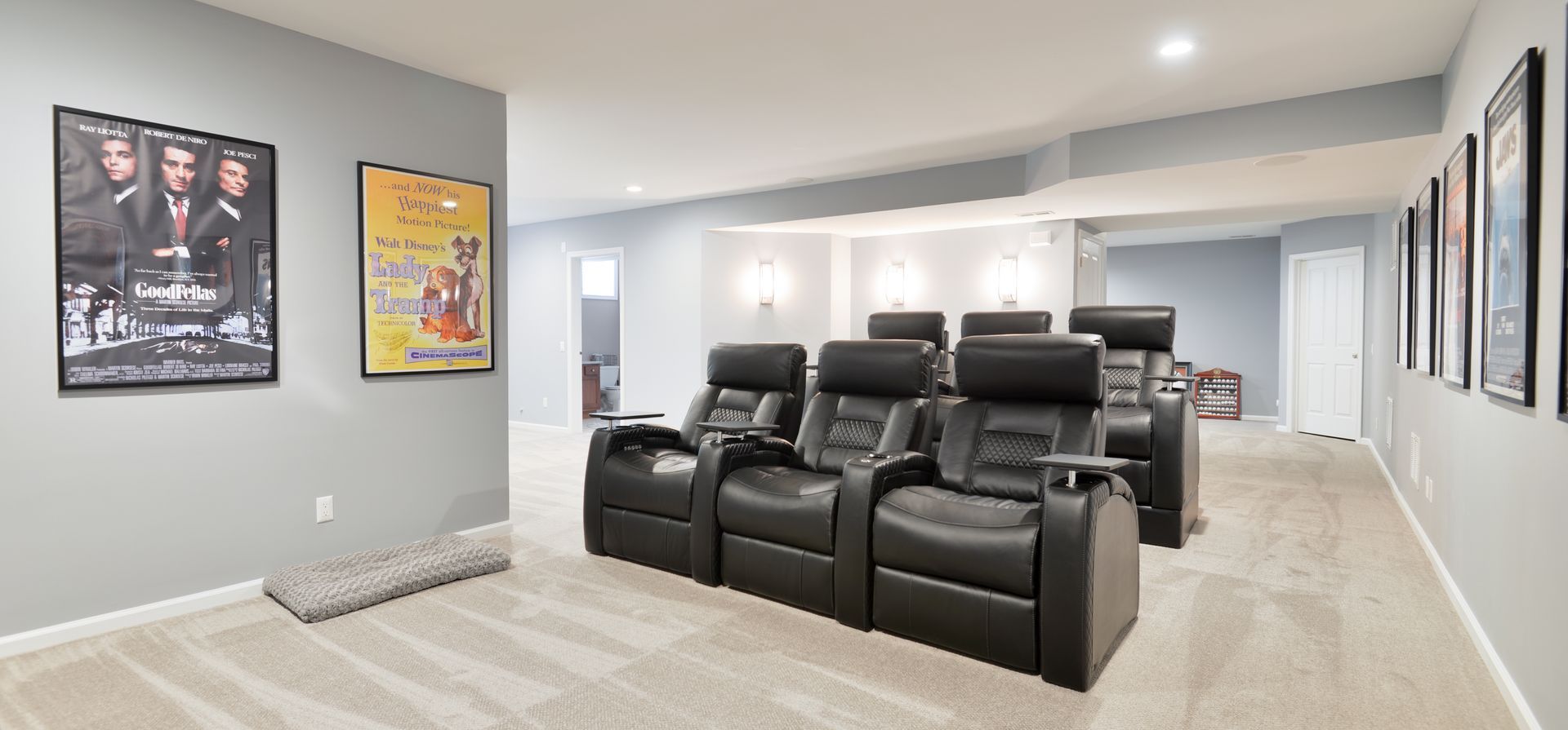 A home theater with four black leather reclining chairs in a row, light carpet, and movie posters on grey walls.