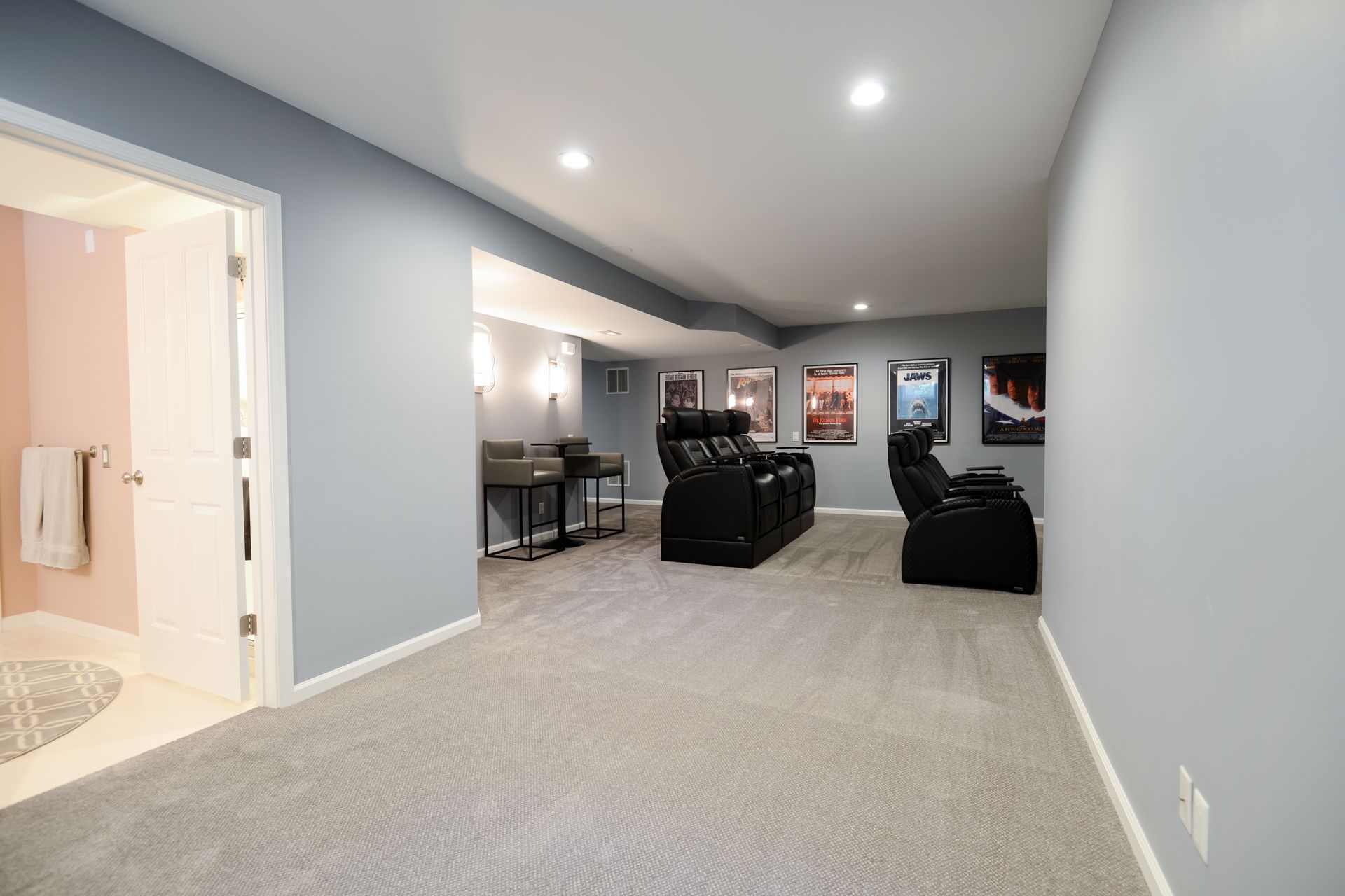 A basement home theater with rows of black leather recliners, movie posters on the walls, and a nearby bathroom entrance.