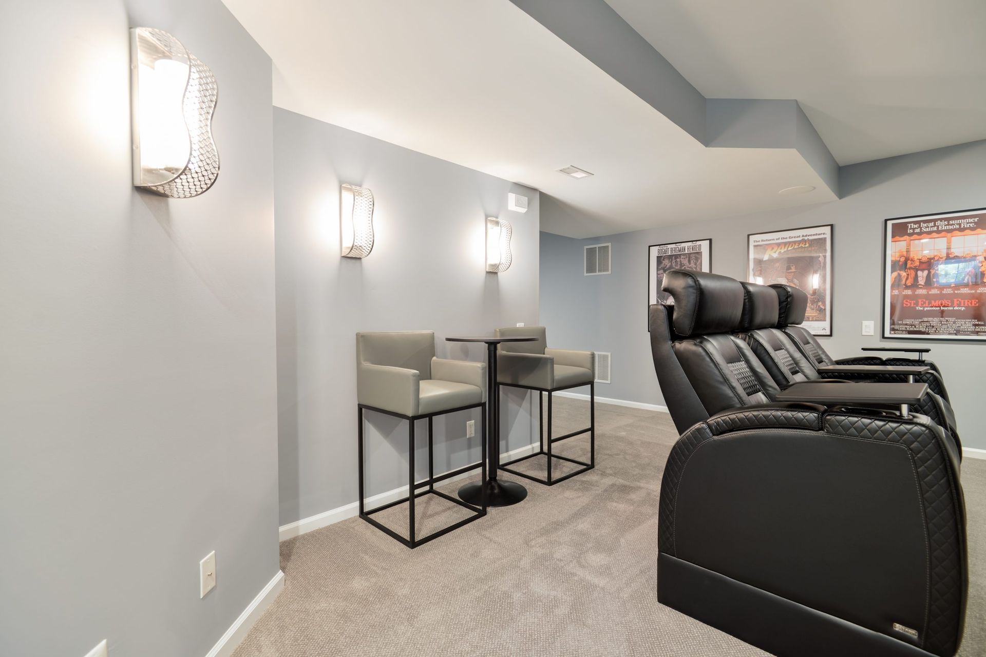 A home theater with grey walls, three black leather recliner seats, and a high-top table with two grey bar stools.