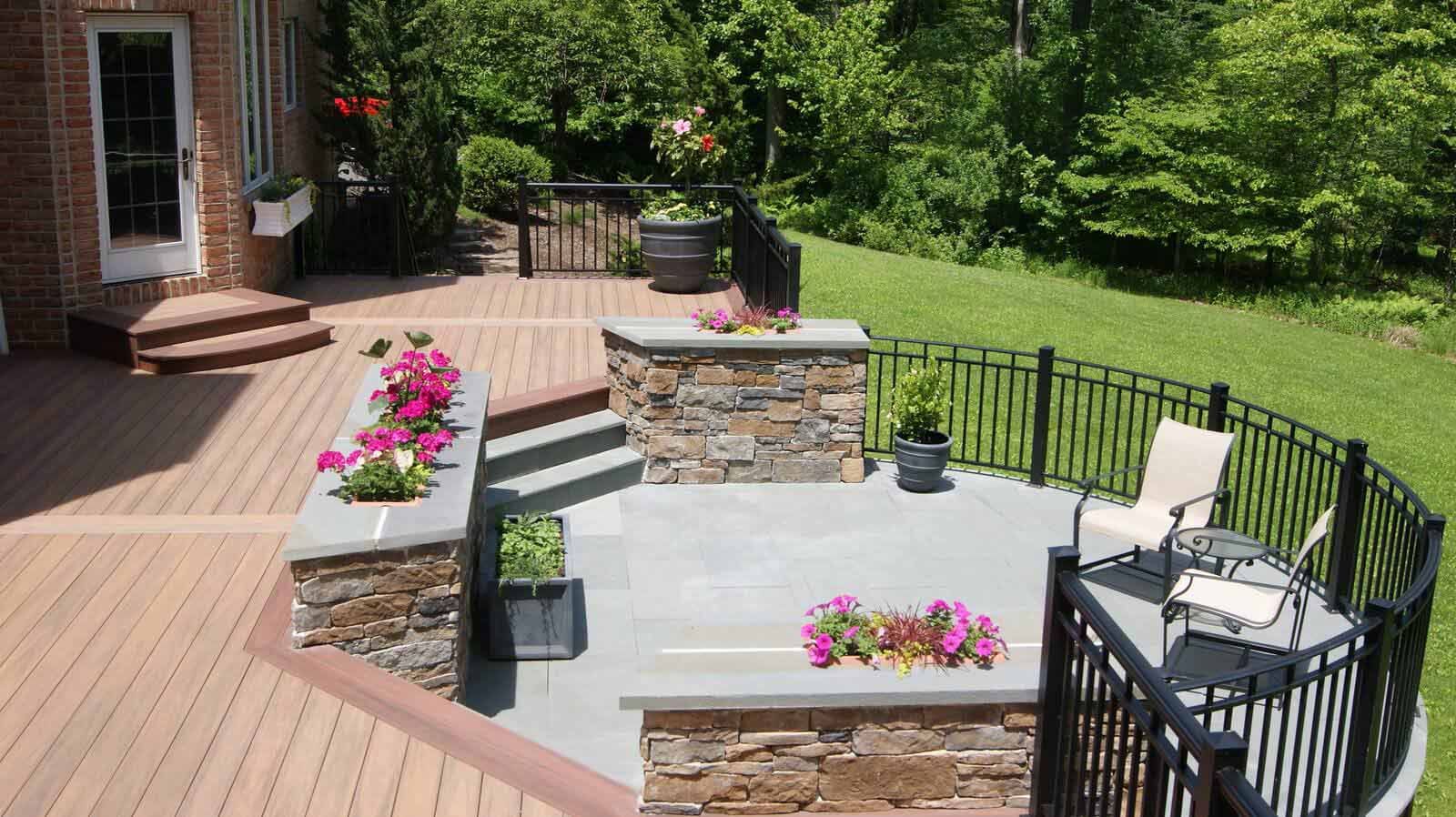 A multilevel backyard patio featuring wood decking, stone retaining walls with flowers, and a curved black metal railing.