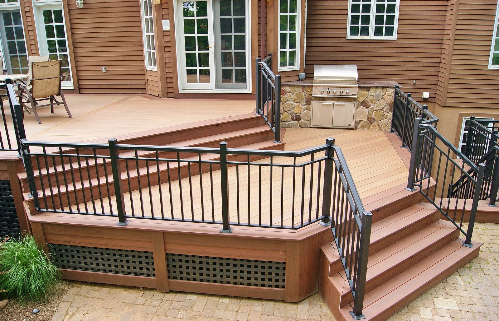 A multi-level wood deck featuring black railings, stairs, and a built-in outdoor grill area against a house wall.