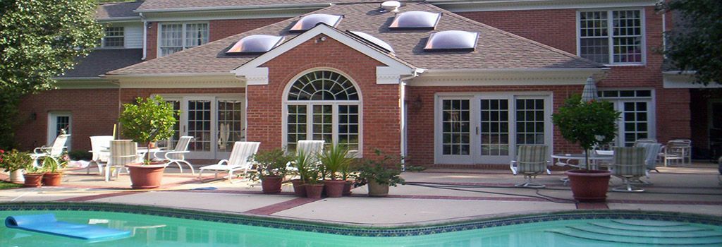 A sunny patio with lounge chairs and potted plants beside a swimming pool, attached to a brick house with large windows.
