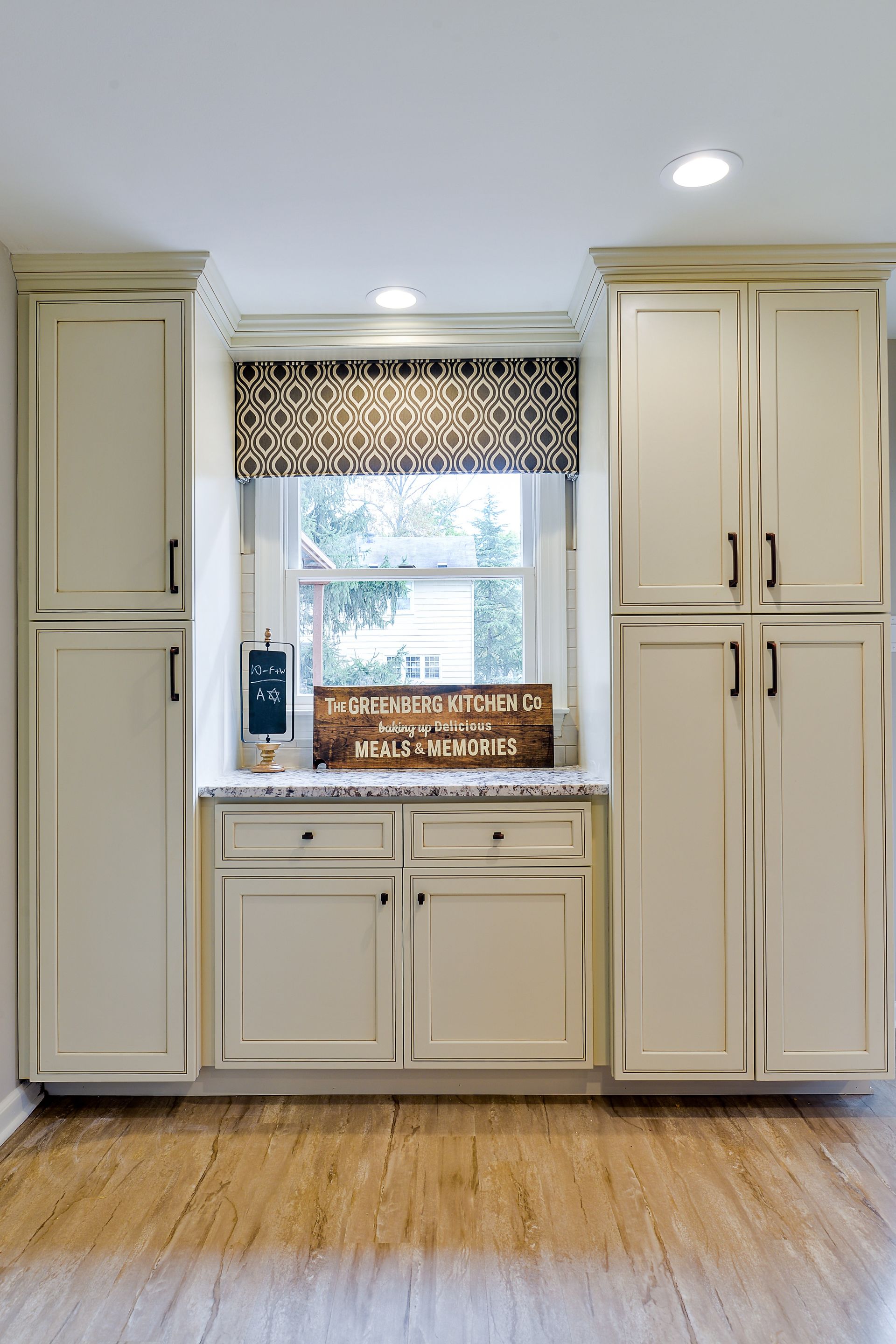 Off-white kitchen cabinets frame a central window with a patterned valance and a small counter with drawers below.